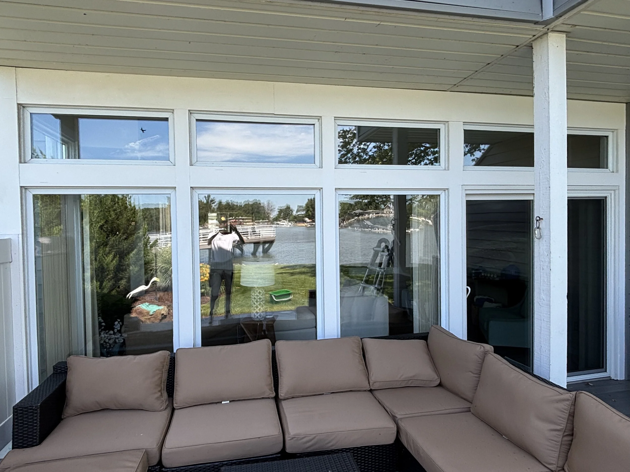 View from a screened porch overlooking a body of water, with a person taking a photo and house interior visible through glass doors and windows, outdoor sofa in the foreground.