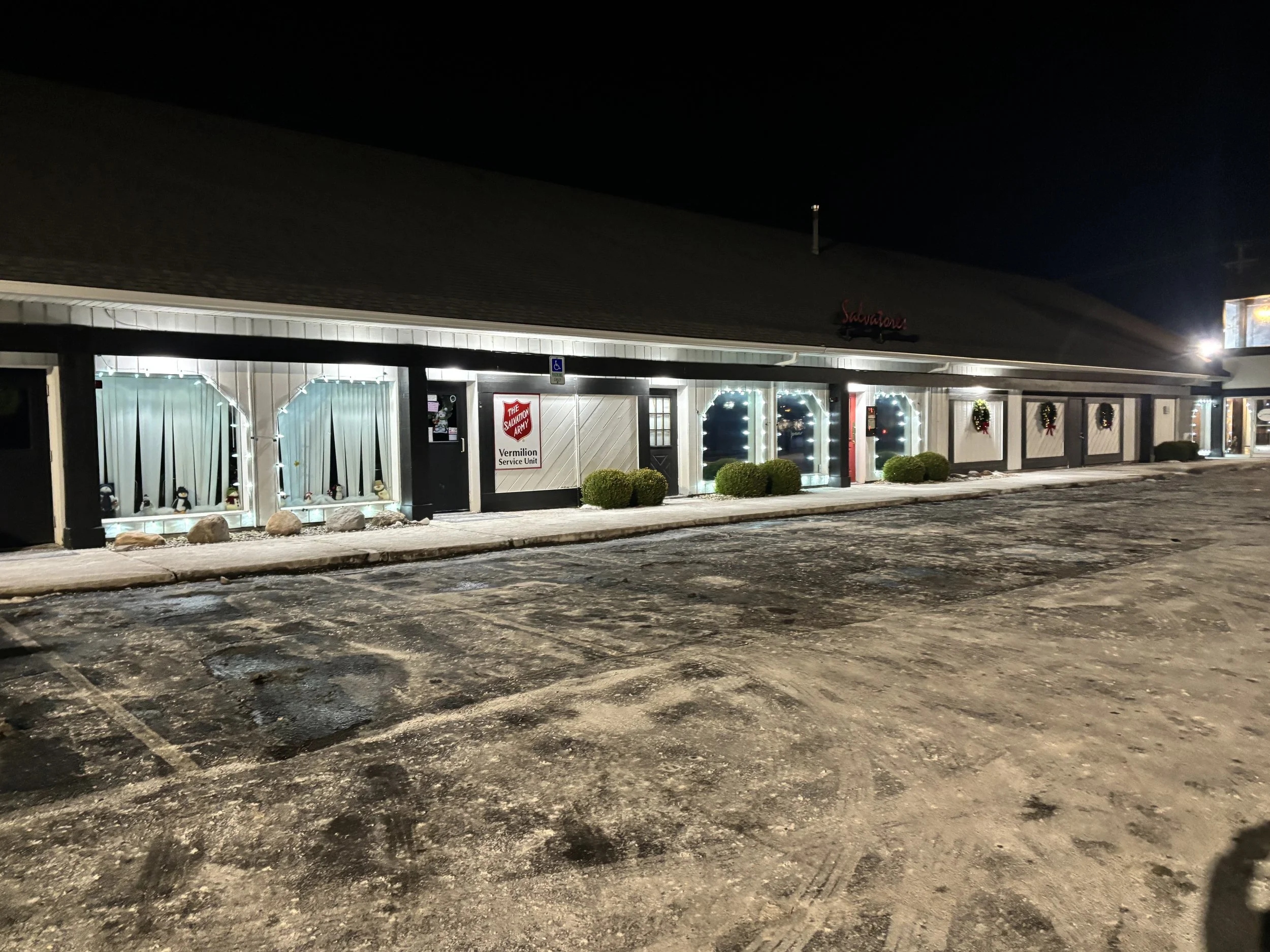Exterior view of a shopping strip at night with holiday decorations, including wreaths on the wall and string lights on windows, and a parking lot in the foreground.