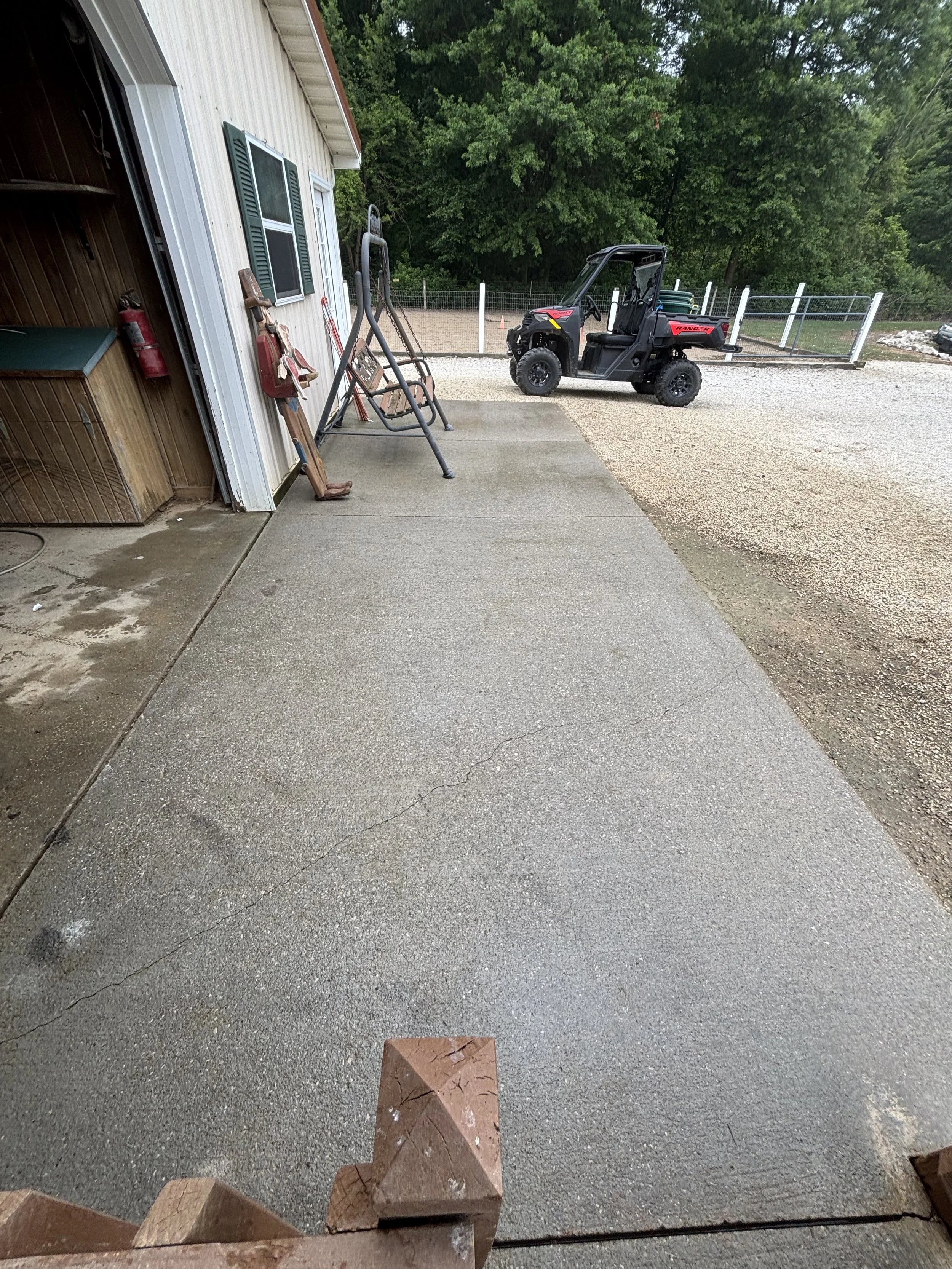 View of a concrete driveway outside a building with gardening tools, a swing set, and a utility vehicle, with trees in the background.