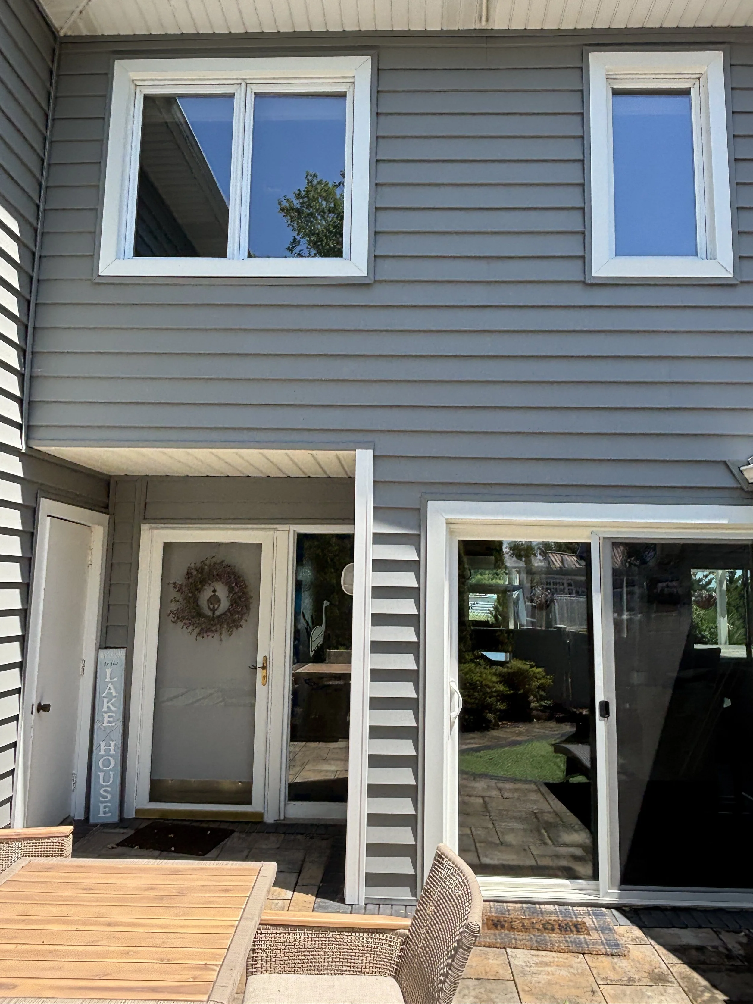 View of a house's back entrance with sliding glass door, a decorative wreath, outdoor seating, and a sign that reads 'Lake House'.
