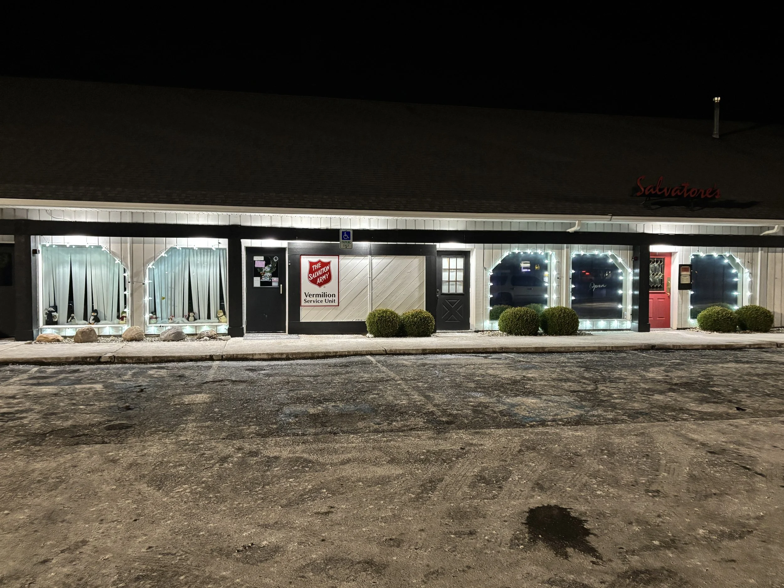 The storefront of a Salvation Army building decorated with Christmas lights, with windows displaying holiday decorations, and a red door, at night.