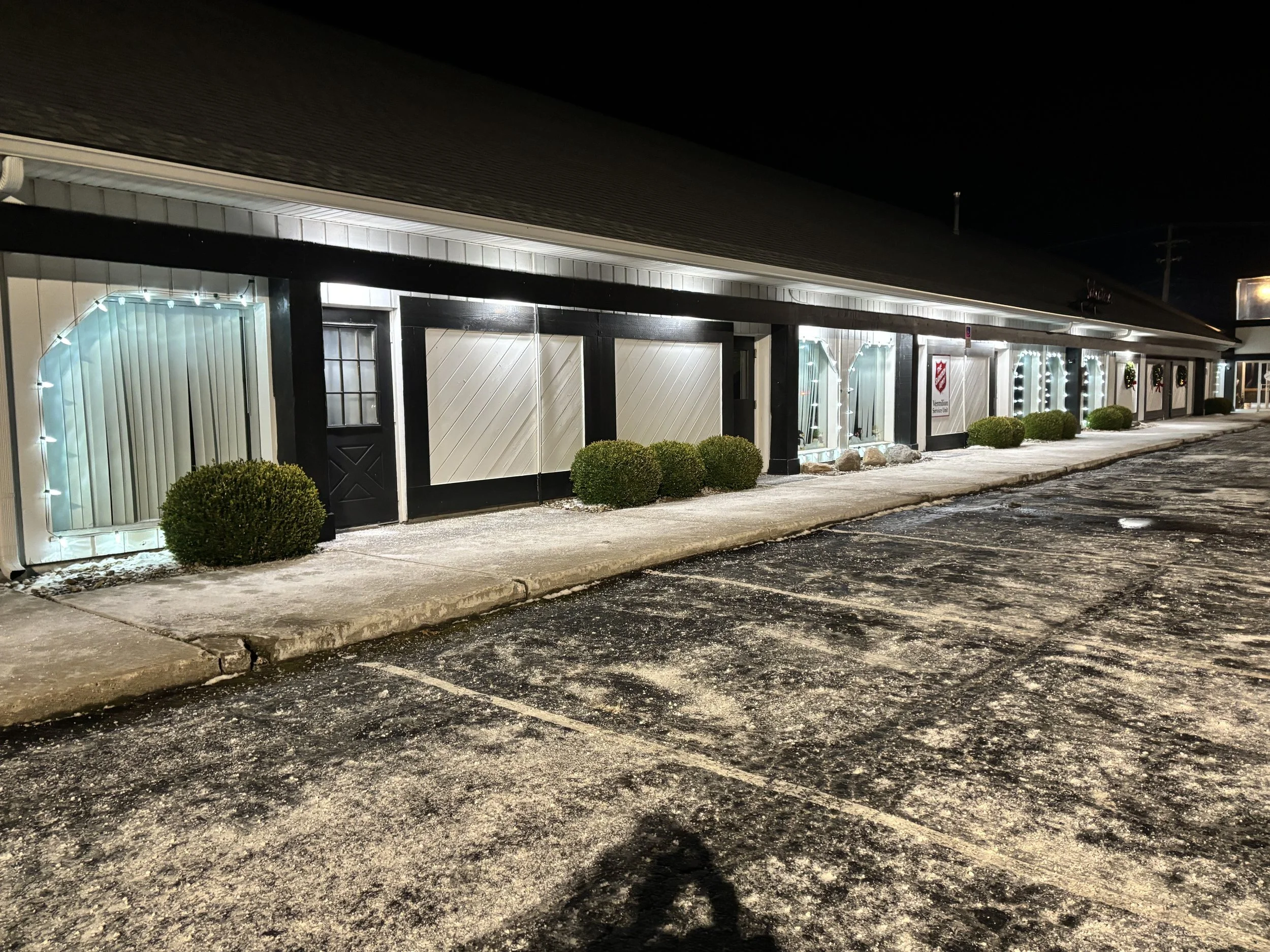 A long building decorated with Christmas lights, black trim, and bushes along the sidewalk at night. The parking lot in the foreground is partially covered with snow and ice.