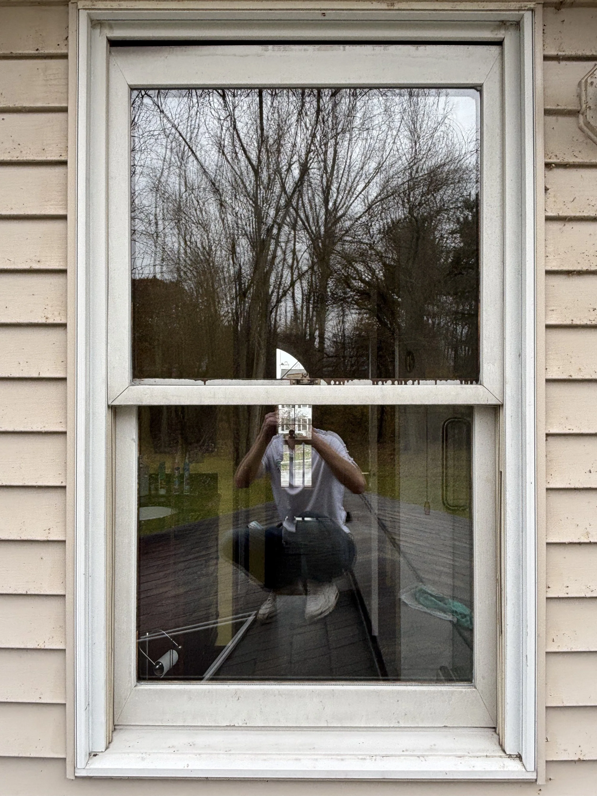 The image shows a window with a reflection of trees and the person taking the photo, who is wearing a white shirt and dark pants, standing outdoors on a deck.
