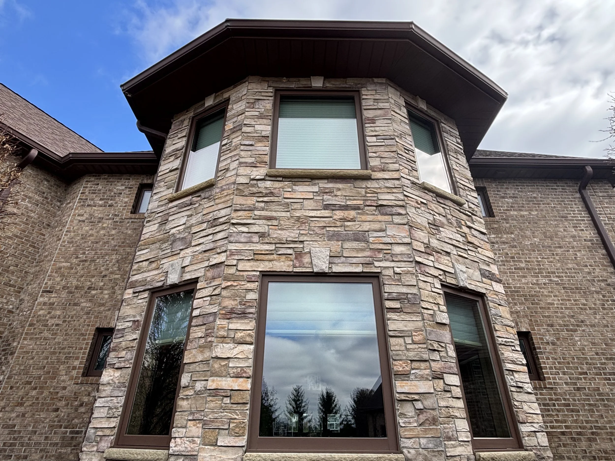 The image shows the exterior of a multi-story brick house with a prominent stone facade section. The house has several windows with brown frames, and the sky above is partly cloudy with patches of blue visible.