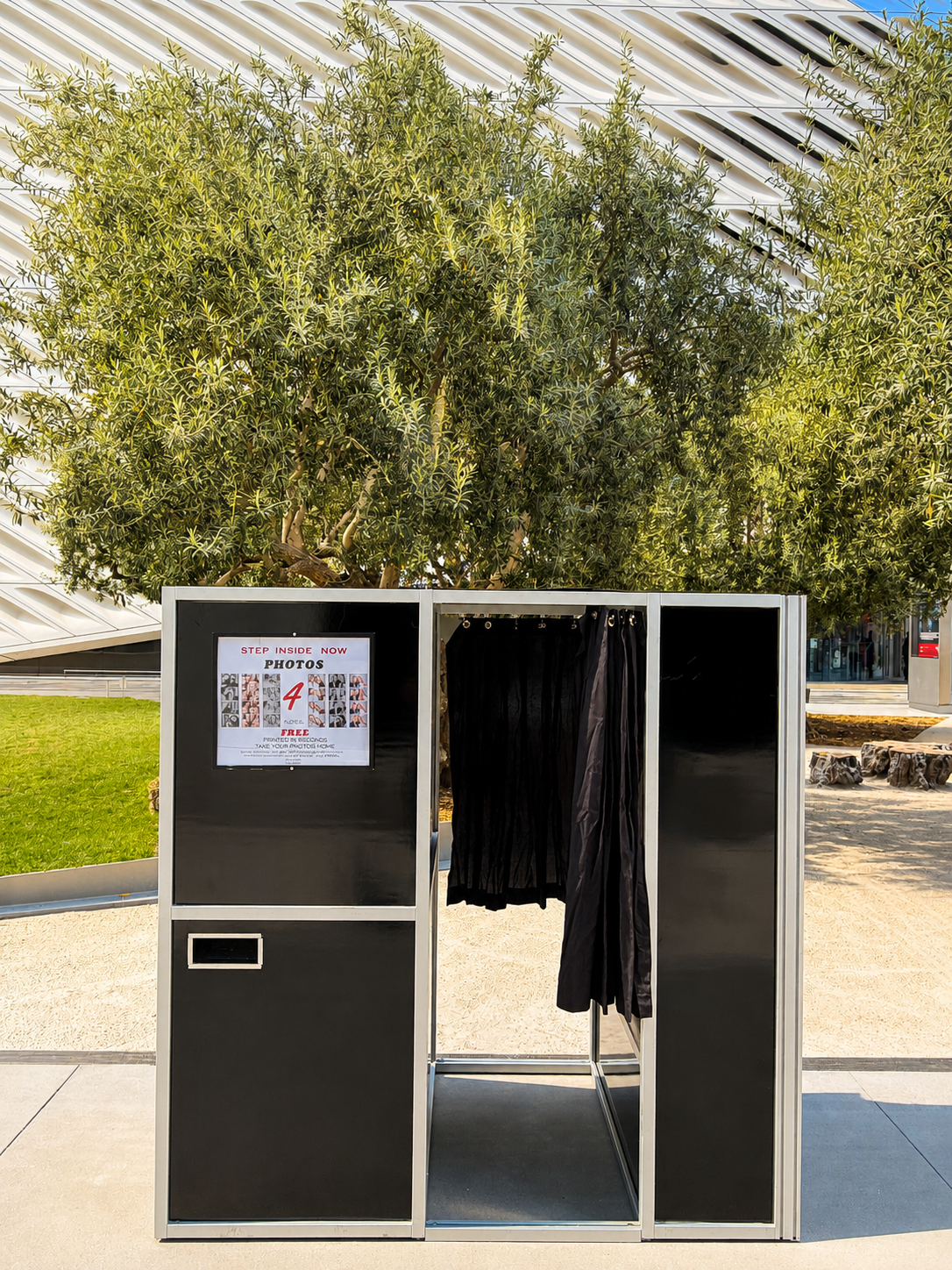 Photo booth with black curtains and frame, sign advertising free photos, outdoor setting with green grass, rocky landscaping, and a large leafy tree in the background, modern building with angled roof.