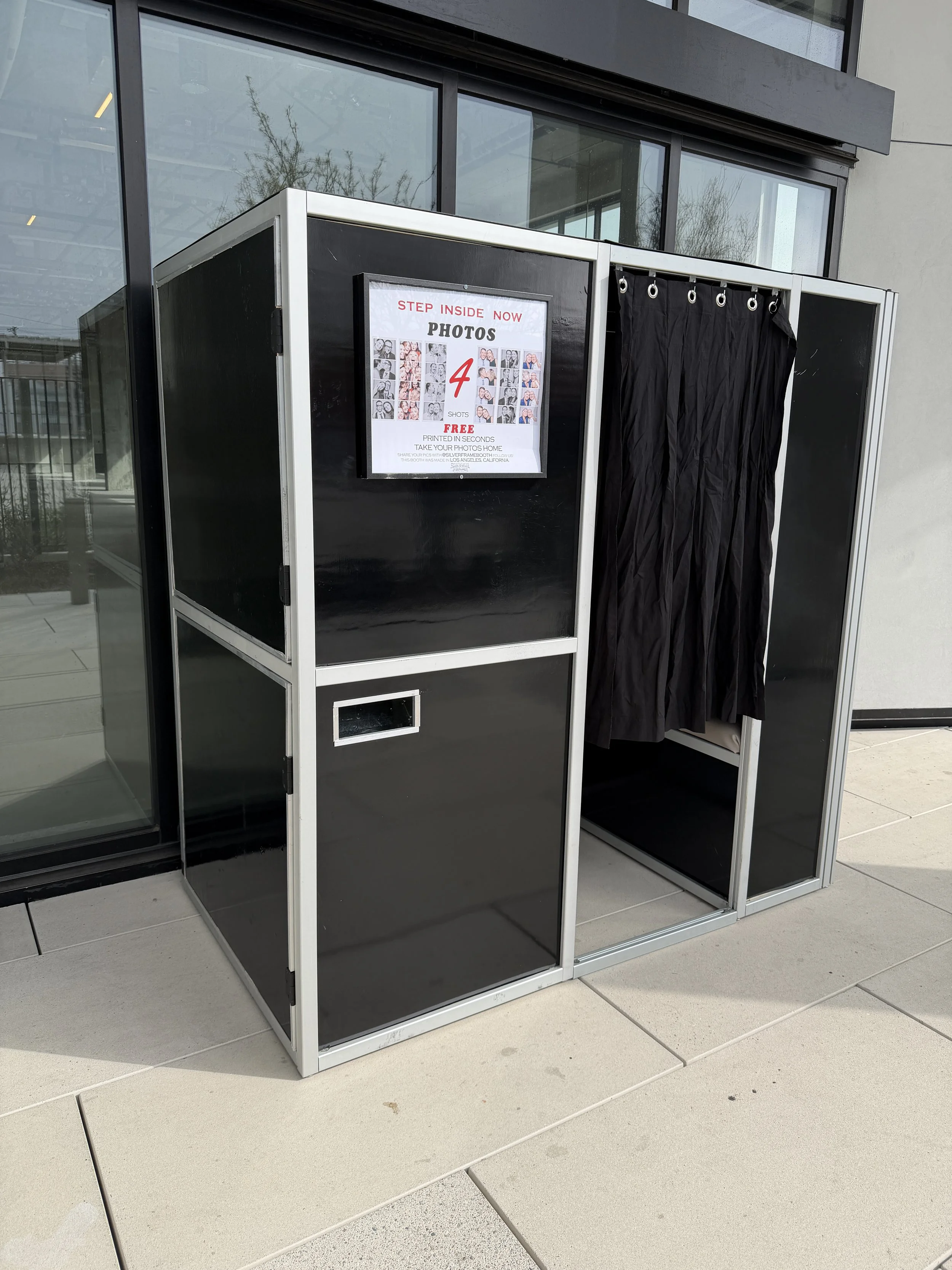 Enclosed photo booth in front of a glass door on cement in a corridor.
