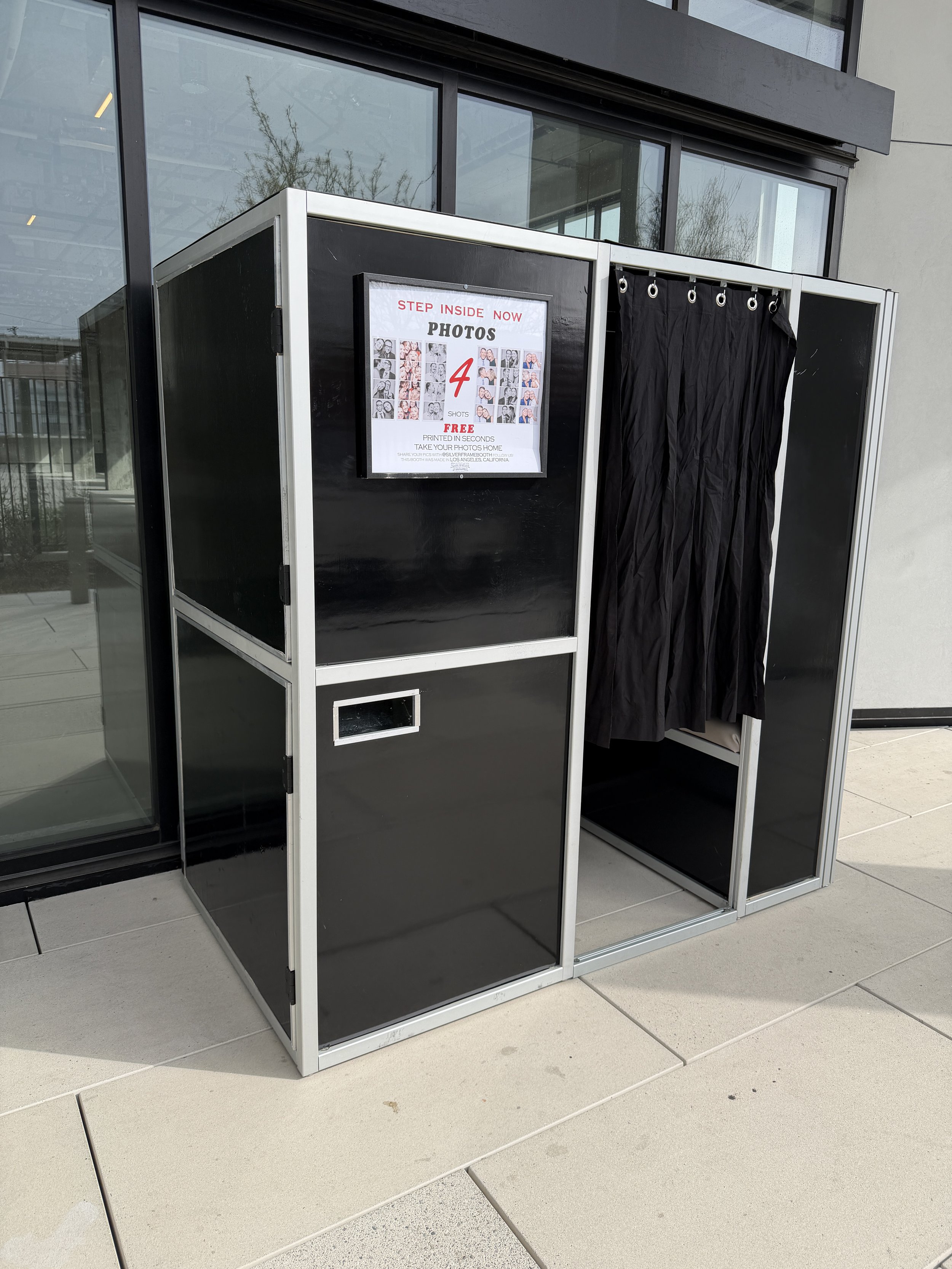 A photo booth with a black curtain, situated outside a building with large glass windows. A sign on the booth advertises free 4-photo strips that are printed in seconds.
