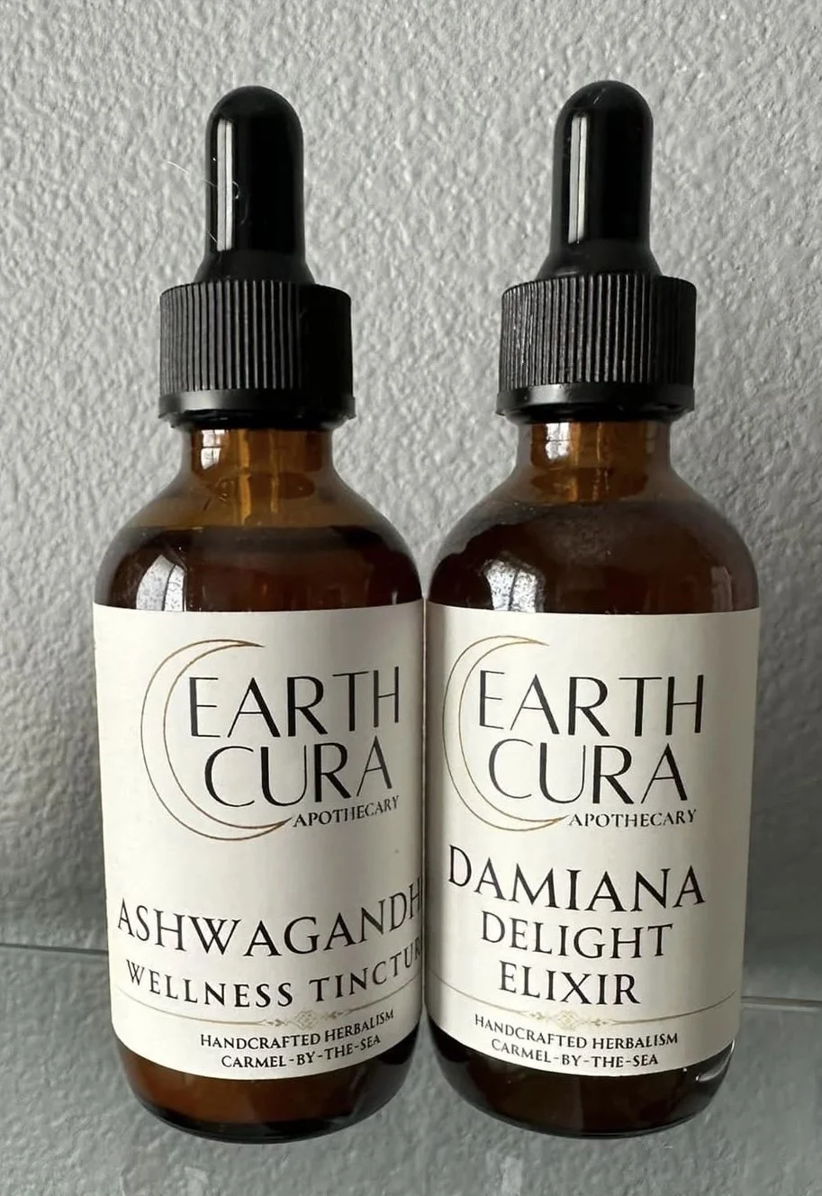 Two brown glass bottles with black dropper caps labeled Earth Cura with herbal remedies, one named Ashwagandha Wellness Tincture and the other Damiana Delight Elixir, placed against a textured wall.