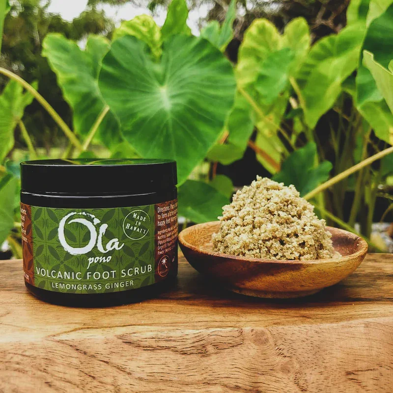 A jar of Ola Pono volcanic foot scrub with lemongrass and ginger next to a wooden bowl filled with the scrub, placed on a wooden surface with green tropical leaves in the background.