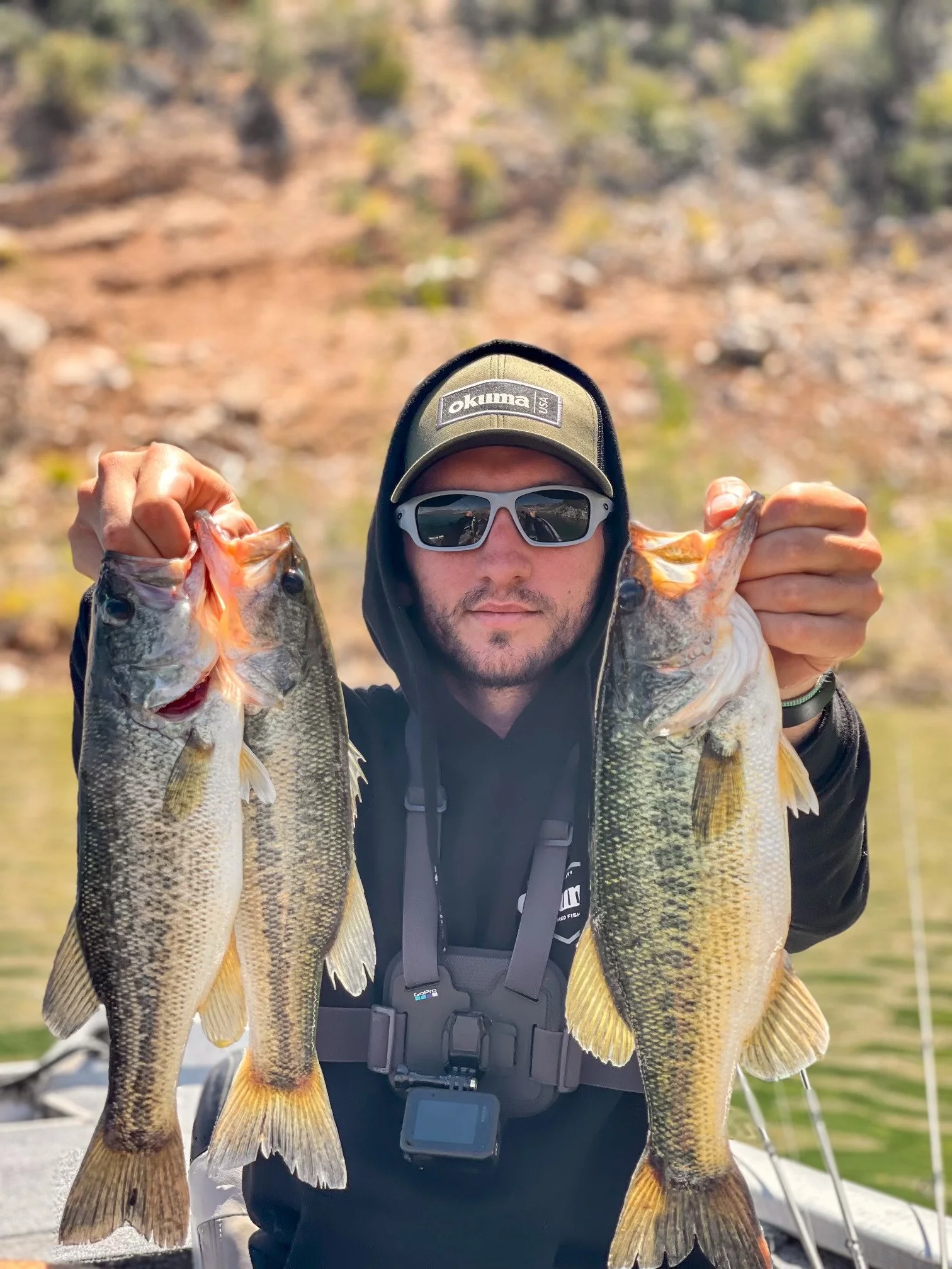 A man wearing sunglasses and a hat holding two large fish, one in each hand, on a boat with a water body and a rocky hill in the background.