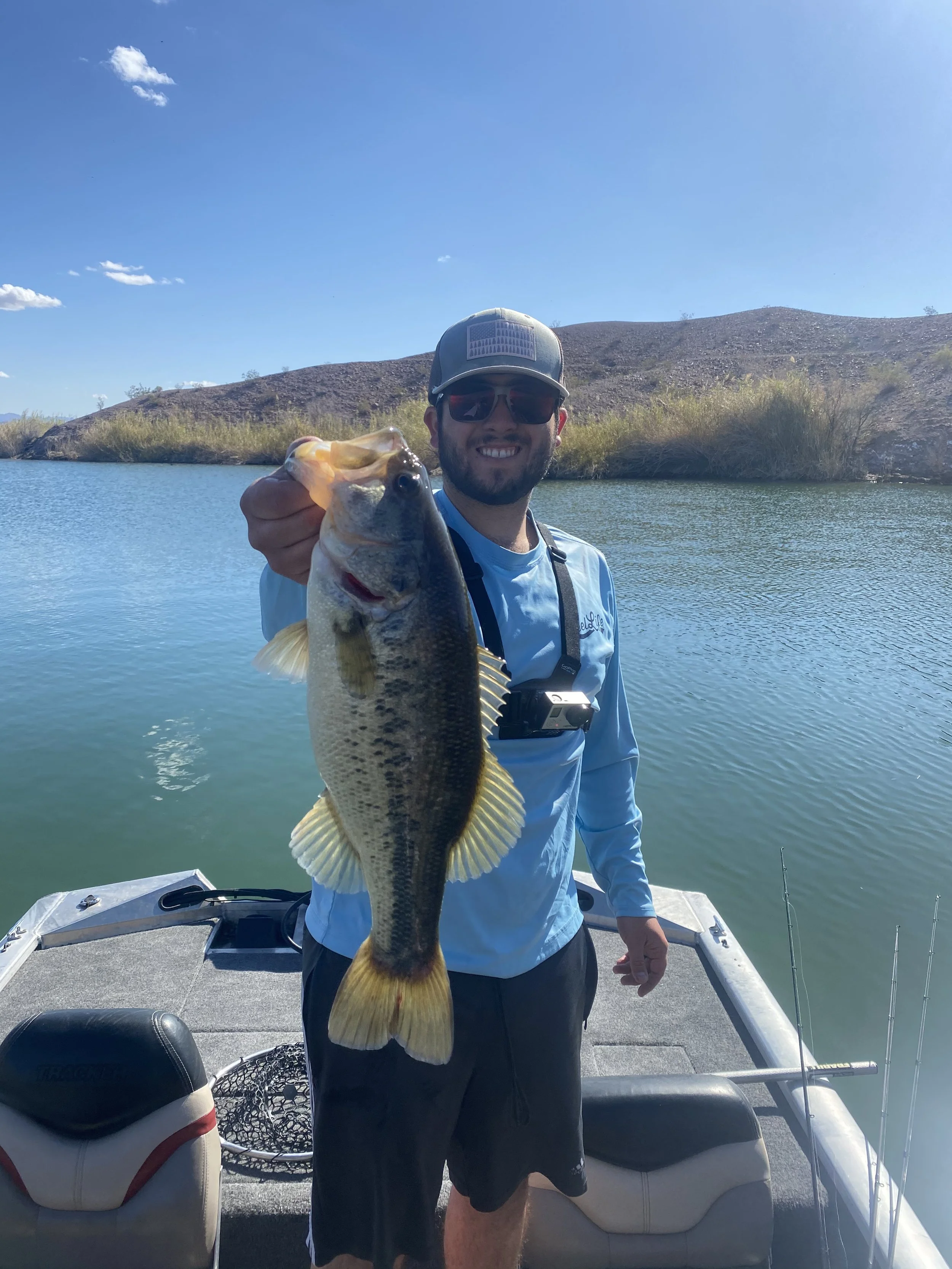 Man on a boat holding a large fish with a wide smile, wearing sunglasses and a baseball cap, with a scenic water and hillside background during daytime.