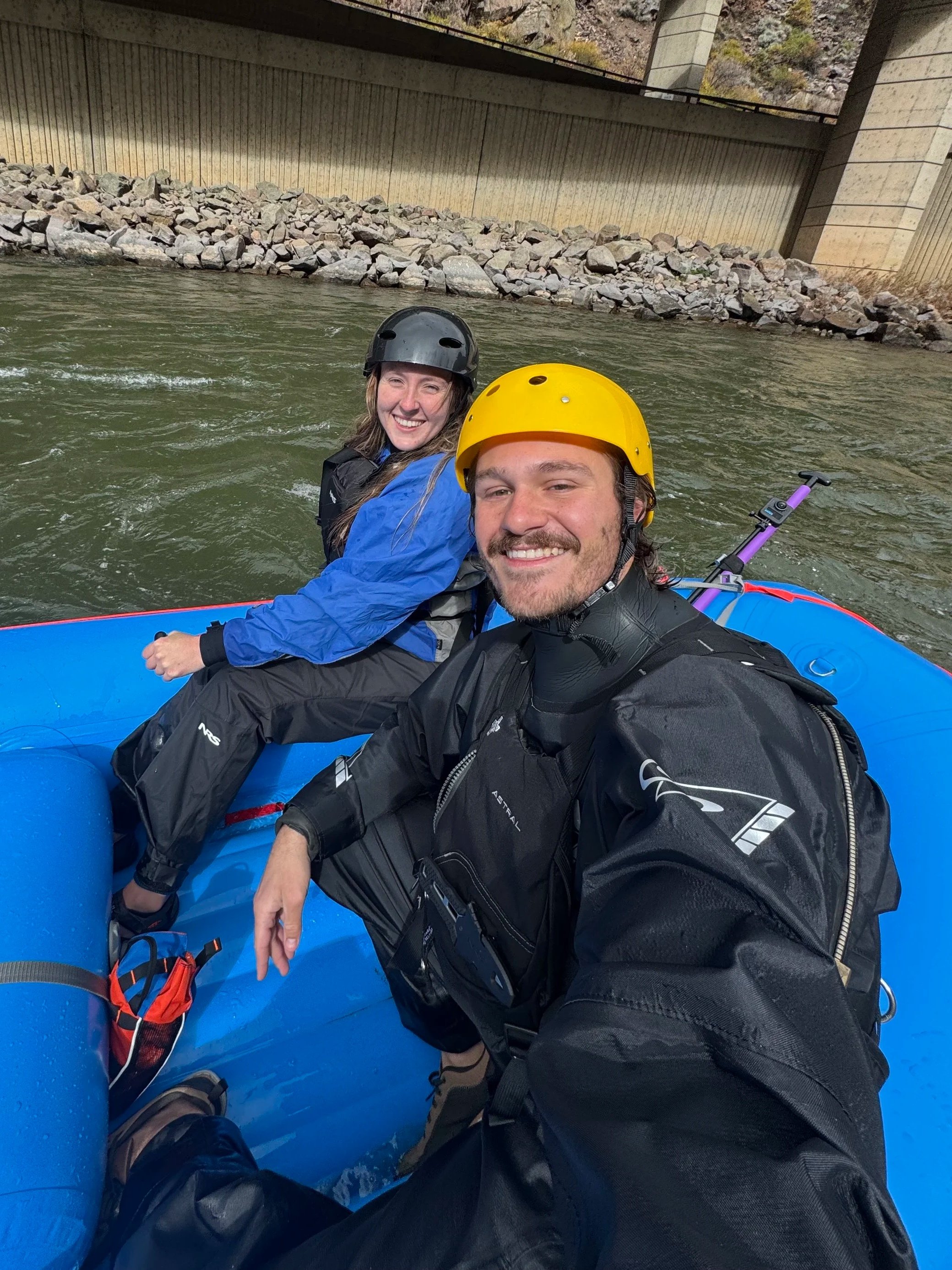 A man and woman in outdoor gear and helmets riding a blue inflatable raft on a river, with a rocky bank and concrete bridge supports in the background.