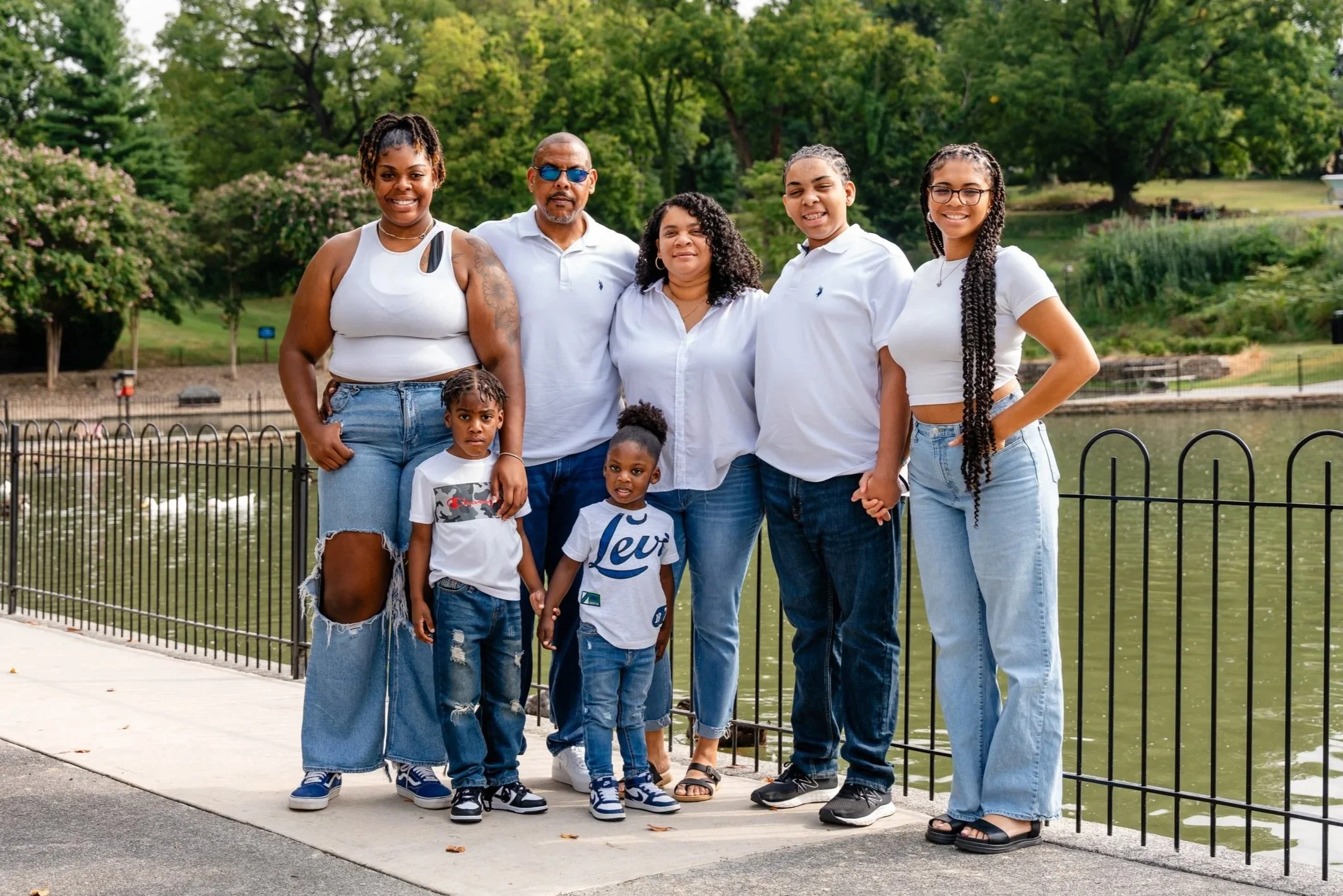 Family photo of seven people, including adults and children, standing outdoors near a water body and fence, with trees in the background.