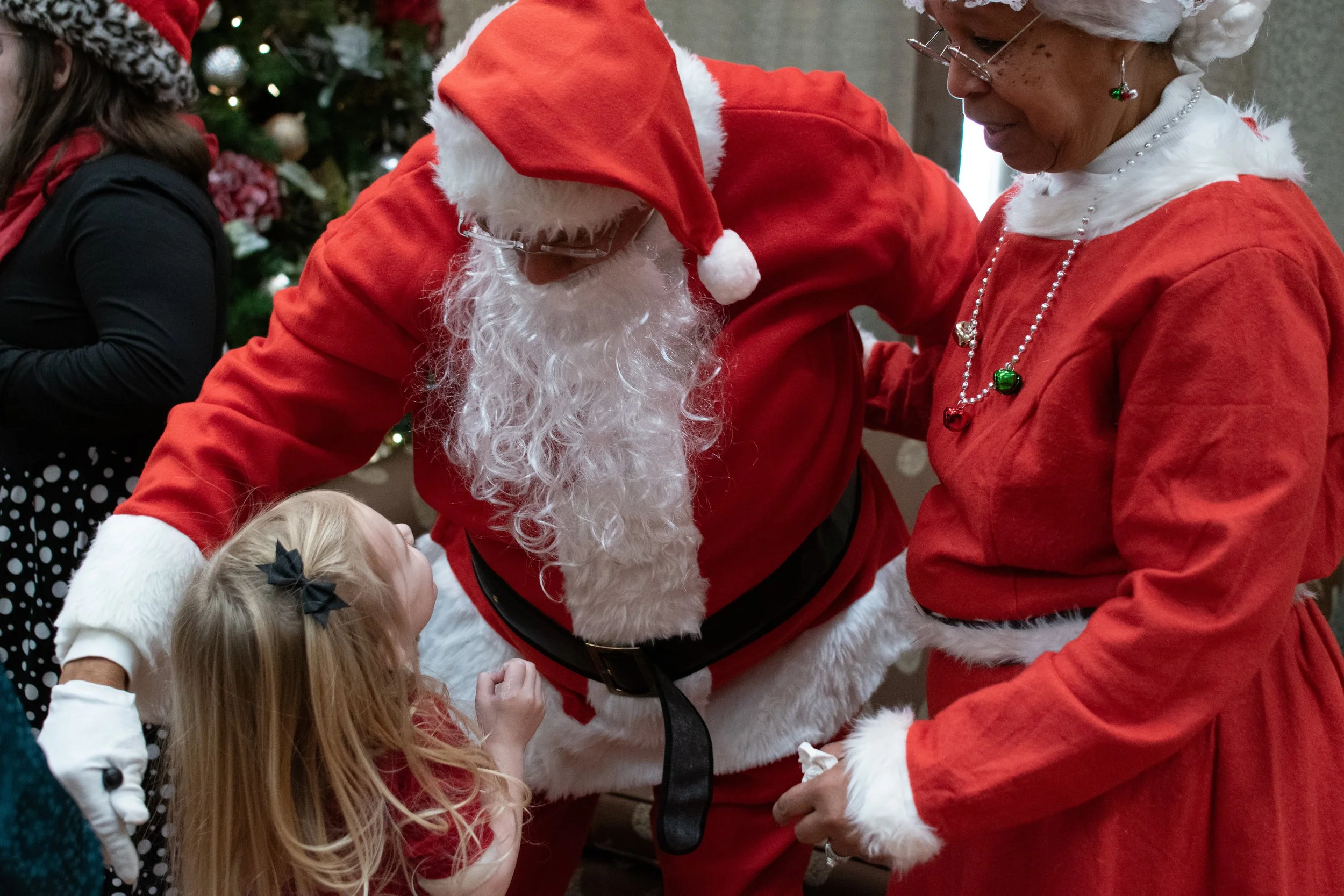 A Christmas scene featuring a man dressed as Santa Claus bending down to greet a young girl with blonde hair and a black bow, while a woman dressed as Mrs. Claus stands nearby. They are indoors with a decorated Christmas tree in the background.