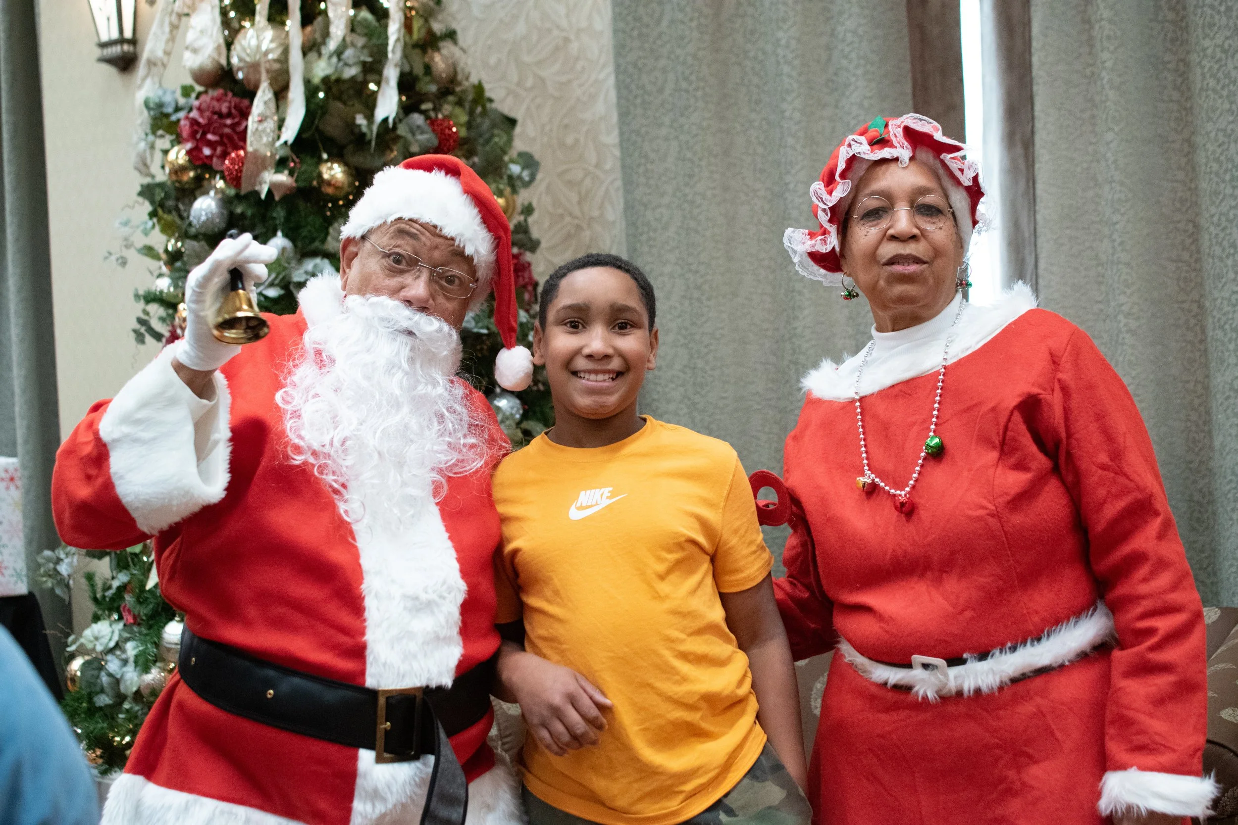A young boy standing between a man dressed as Santa Claus and a woman dressed as Mrs. Claus, all smiling in front of a decorated Christmas tree.