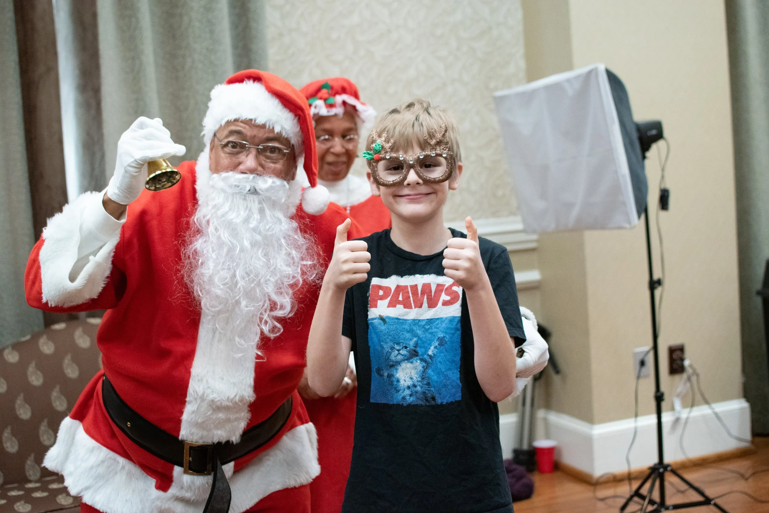 A young boy wearing Christmas glasses and a 'Paws' t-shirt giving two thumbs up next to two adults dressed as Santa and Mrs. Claus, in a room decorated for a holiday event.