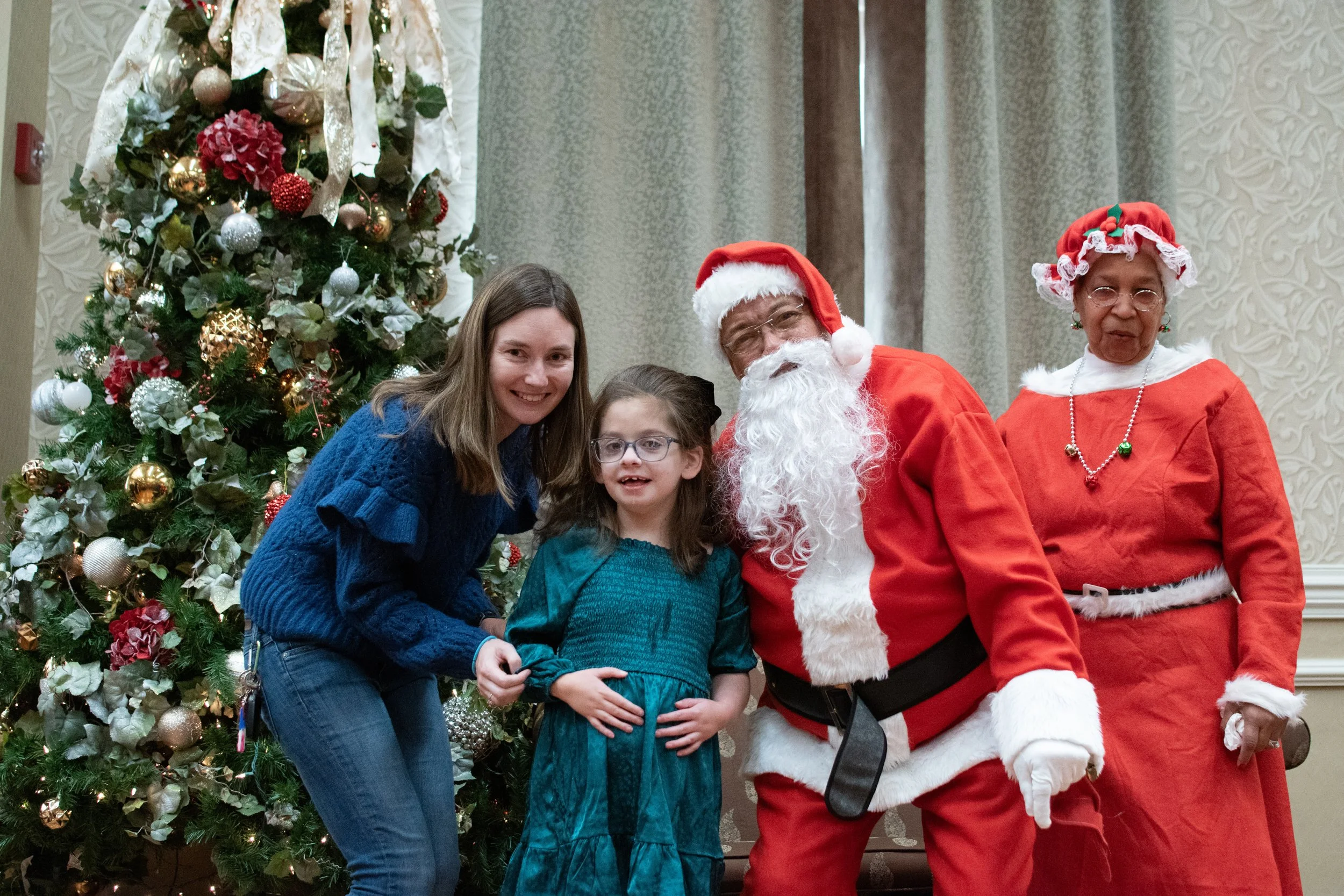 Four people, including a girl, woman, and two adults dressed as Santa Claus and Mrs. Claus, posing in front of a decorated Christmas tree.
