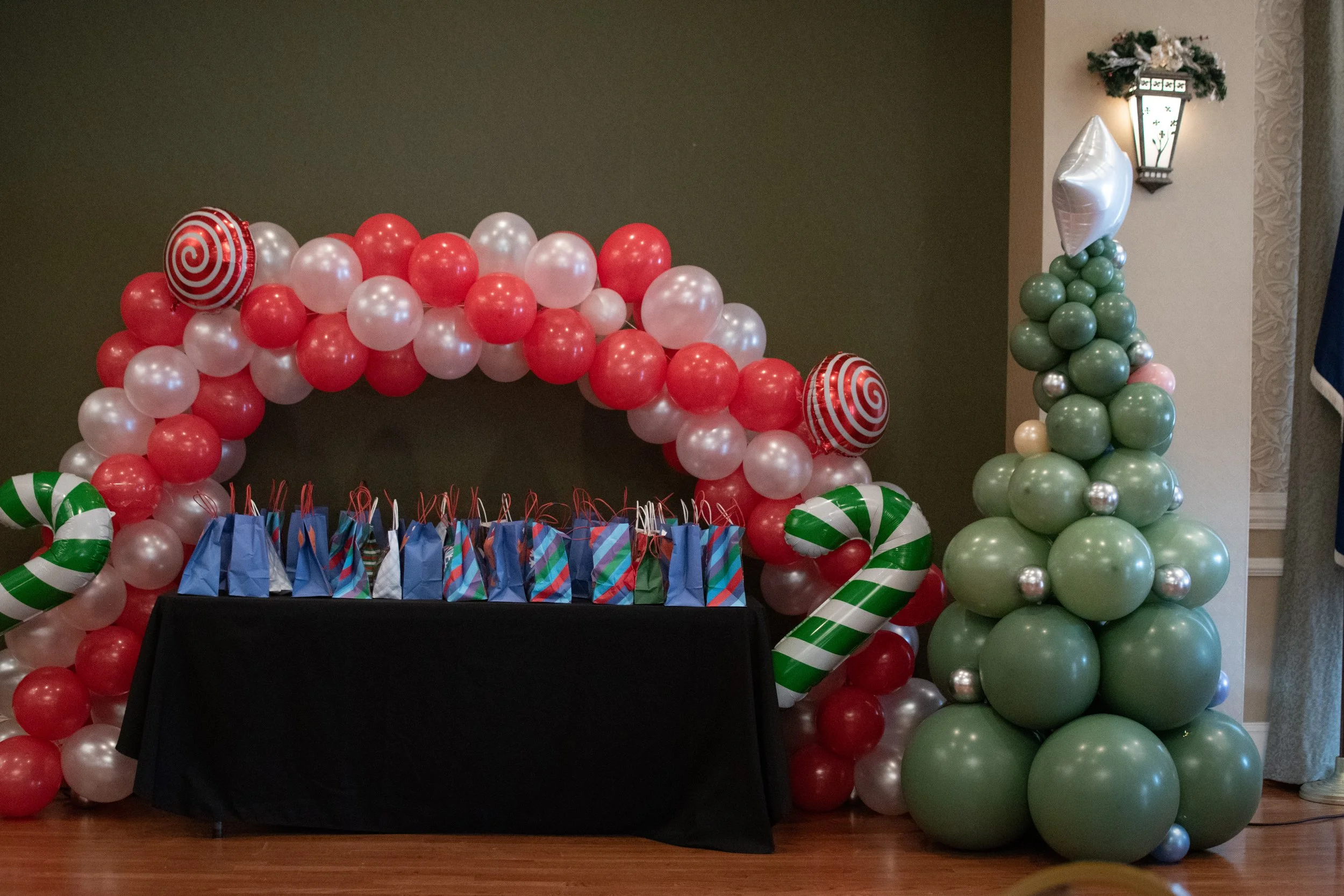 Decorative Christmas balloon arrangement with red and white balloons forming an arch, green and white striped candy cane balloons, a Christmas tree made of green balloons with silver ornaments, and presents on a black-covered table in an indoor setting.
