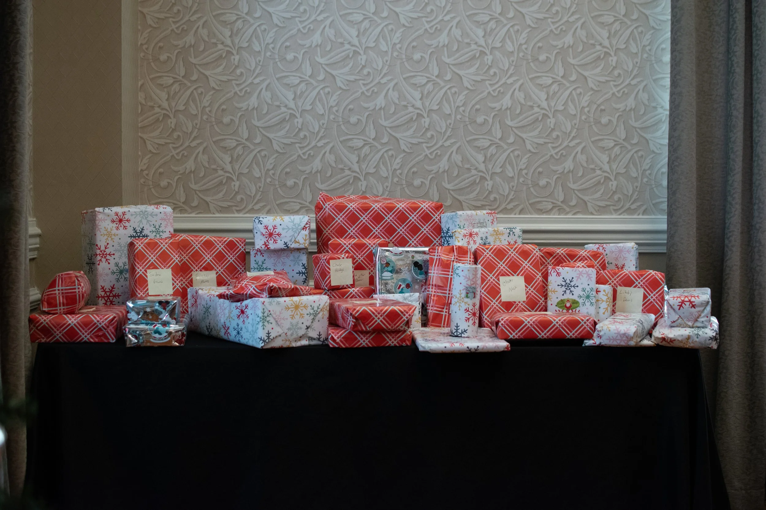 Table with numerous wrapped Christmas presents in festive paper, arranged against a wall with ornate wallpaper and curtains.