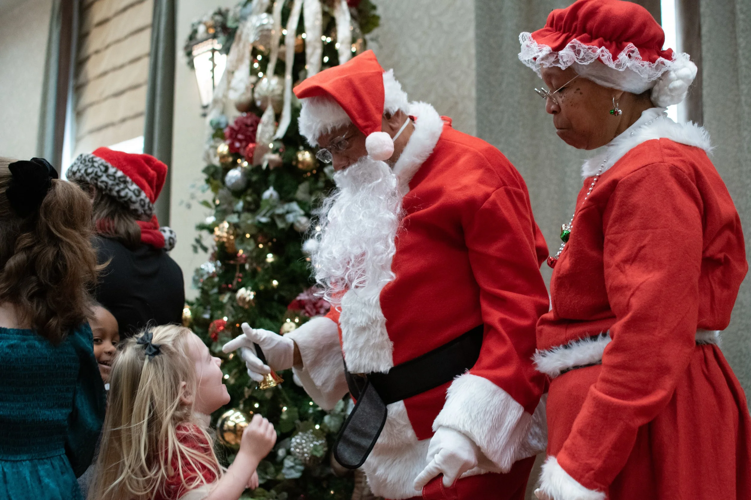 Santa Claus in a red suit with white trim, handing a bell to a young girl in front of a decorated Christmas tree, with a woman dressed as Mrs. Claus standing beside him, both in red outfits with white fur trim, during a holiday event.