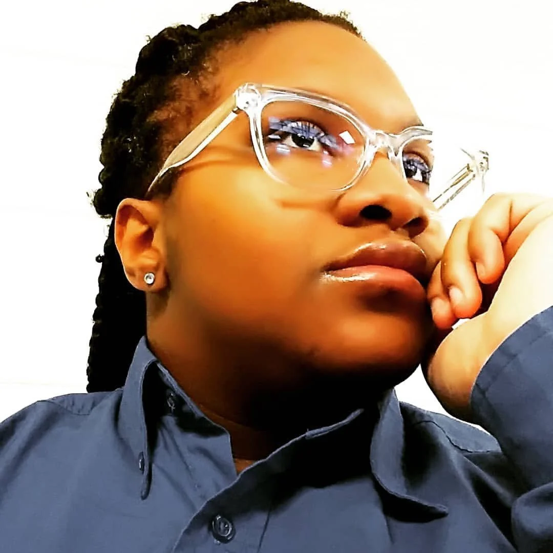 Close-up of a woman wearing clear glasses and a dark blue shirt, resting her chin on her hand and looking thoughtfully to the side.