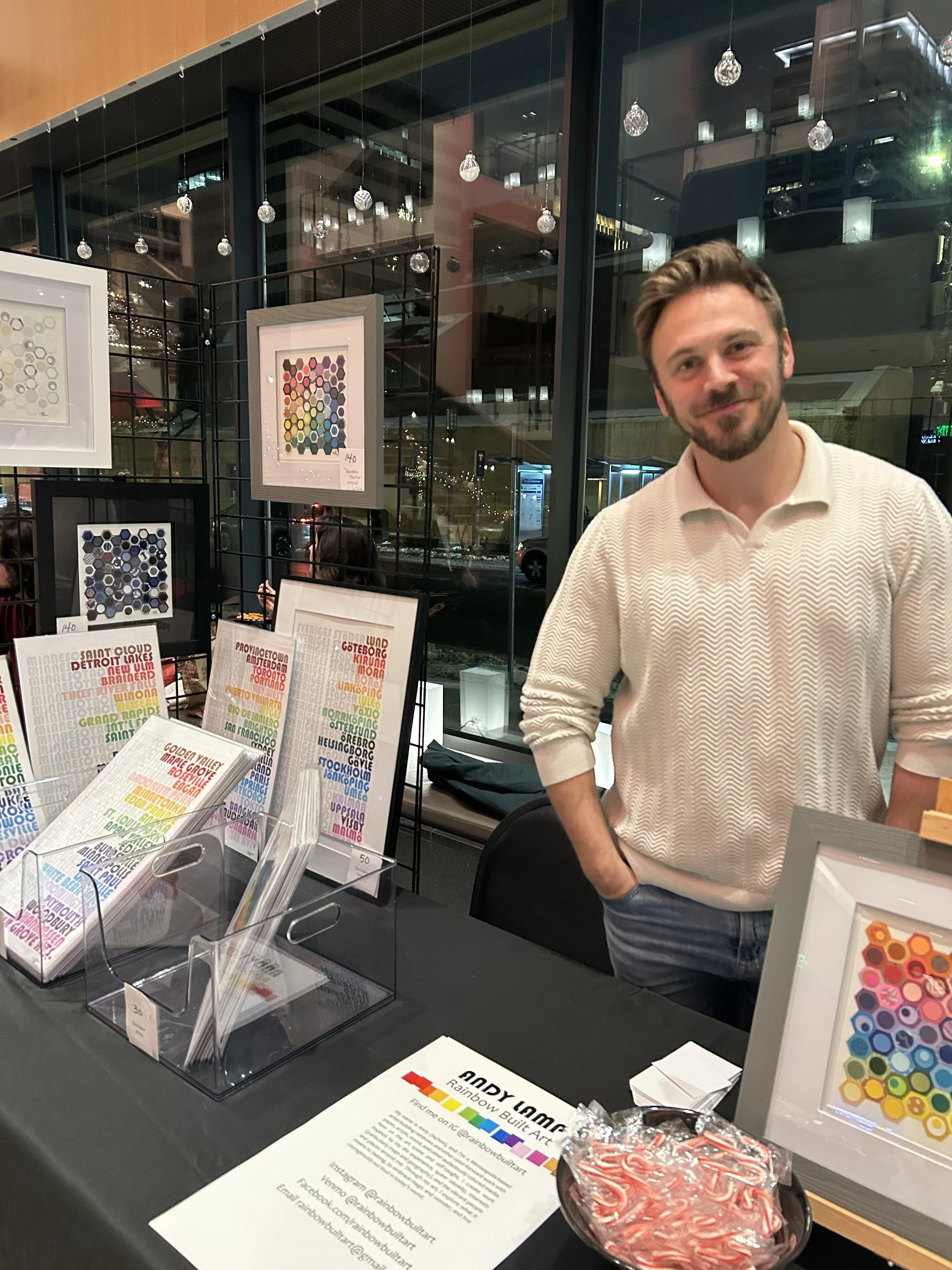 A man with a beard and short hair standing behind a table at an art exhibition, smiling at the camera. The table displays colorful artwork and printed materials, including a rainbow-colored art piece and informational flyers. In the background, there are framed artworks on display, large windows with reflections, and hanging decorative objects.