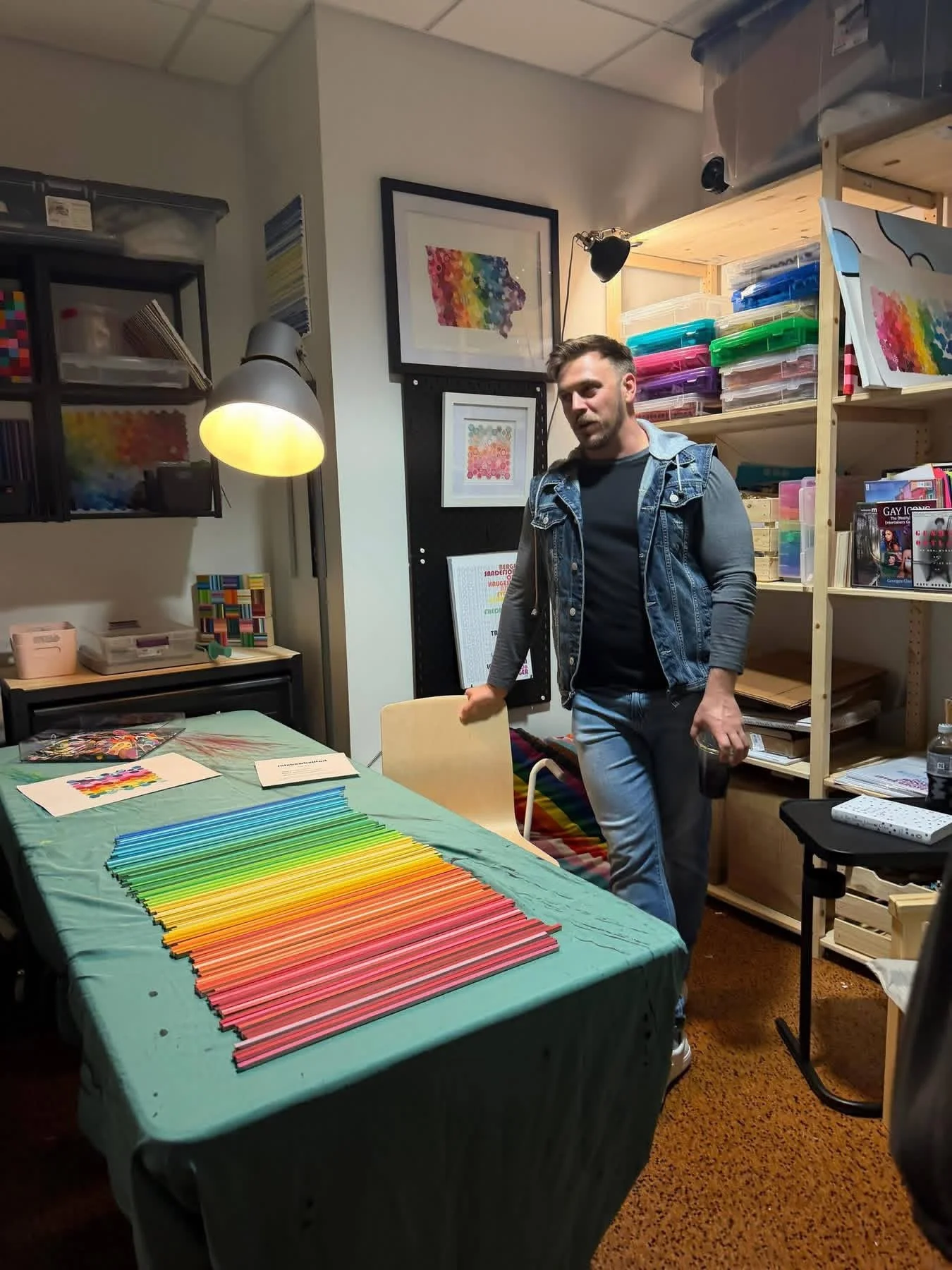 A man with short dark hair, wearing a denim jacket and jeans, in a room filled with rainbow-colored art supplies and artwork. He is standing next to a table covered with rainbow-colored paper strips and smiling slightly. The room has shelves with colorful storage boxes, framed rainbow artwork on the wall, and a black floor lamp.