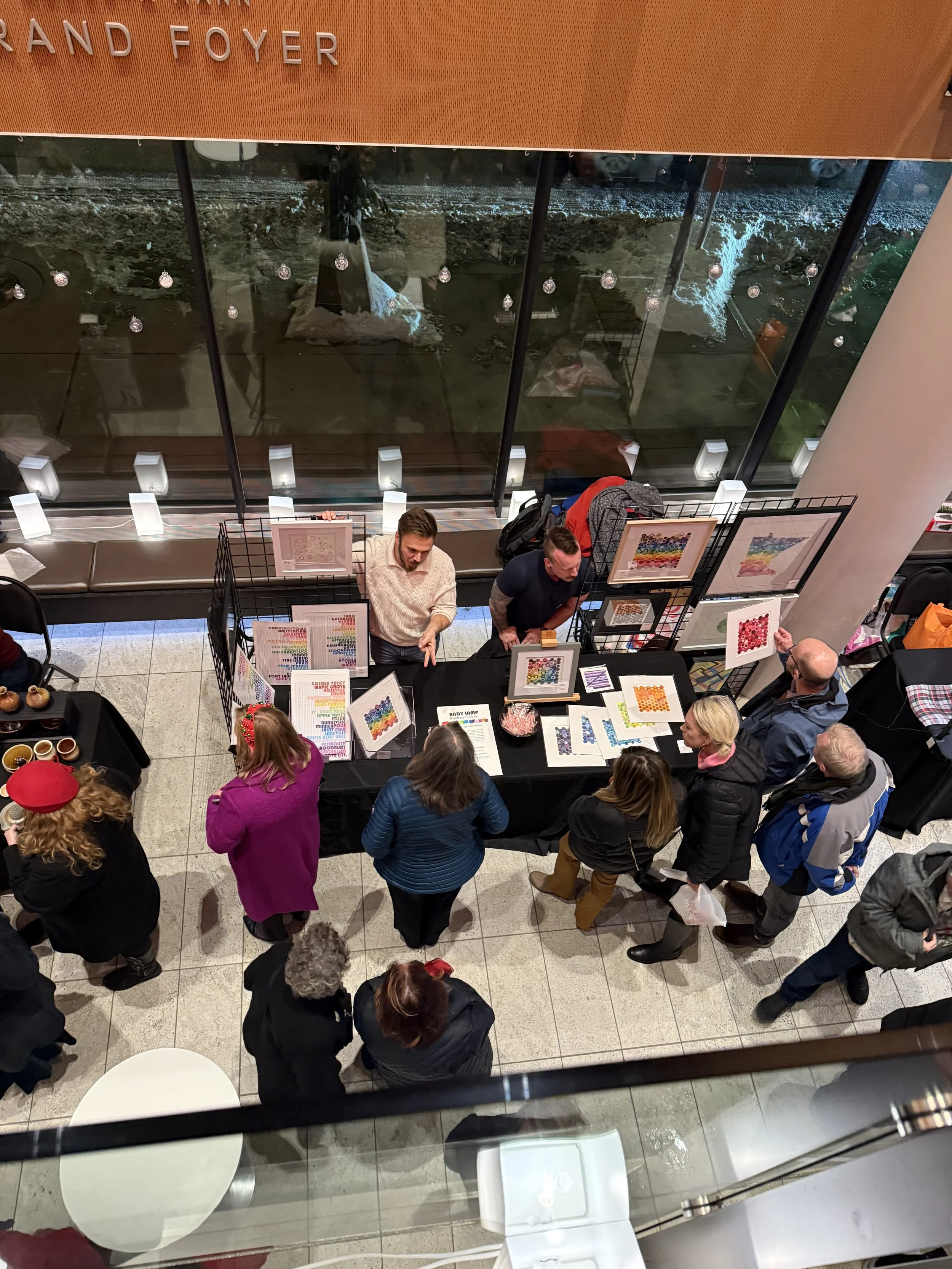 People gathered around a display table at an indoor event, viewing colorful artwork and prints. A large window behind the table shows a night view of a park or landscape outside.