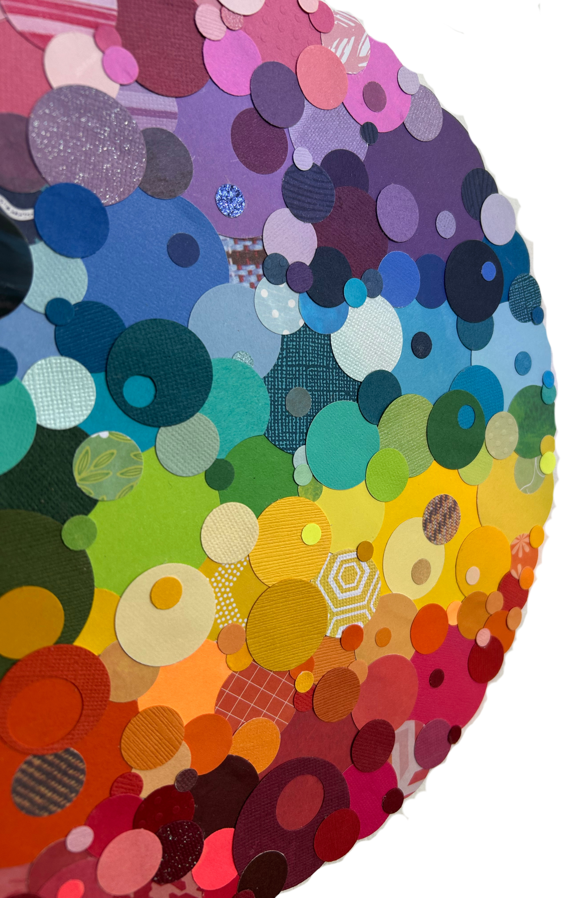A colorful circular art piece made from numerous layered paper circles of various sizes and colors, arranged in a gradient from red and orange in the bottom left to purple and pink at the top right.