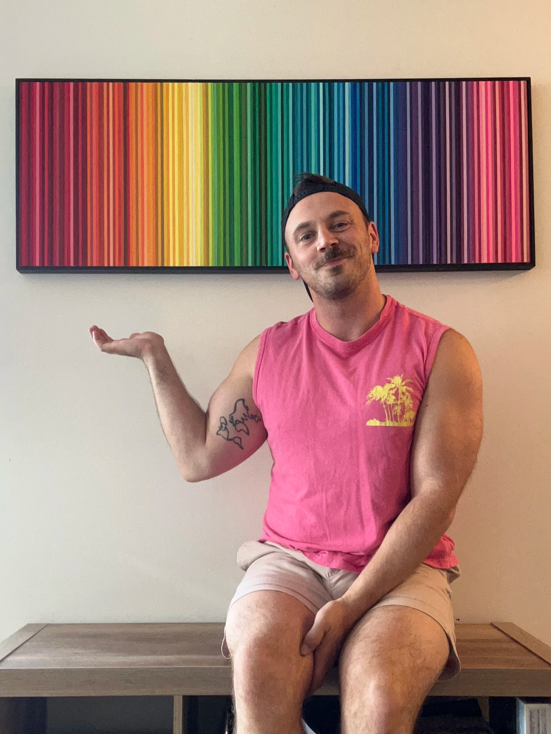 A man with a beard and tattoos on his arm, wearing a pink sleeveless shirt and beige shorts, is sitting on a wooden bench next to a wall. He is smiling and gesturing with his left hand while looking at the camera. Behind him, there is a colorful rectangular artwork featuring vertical stripes in a rainbow gradient.