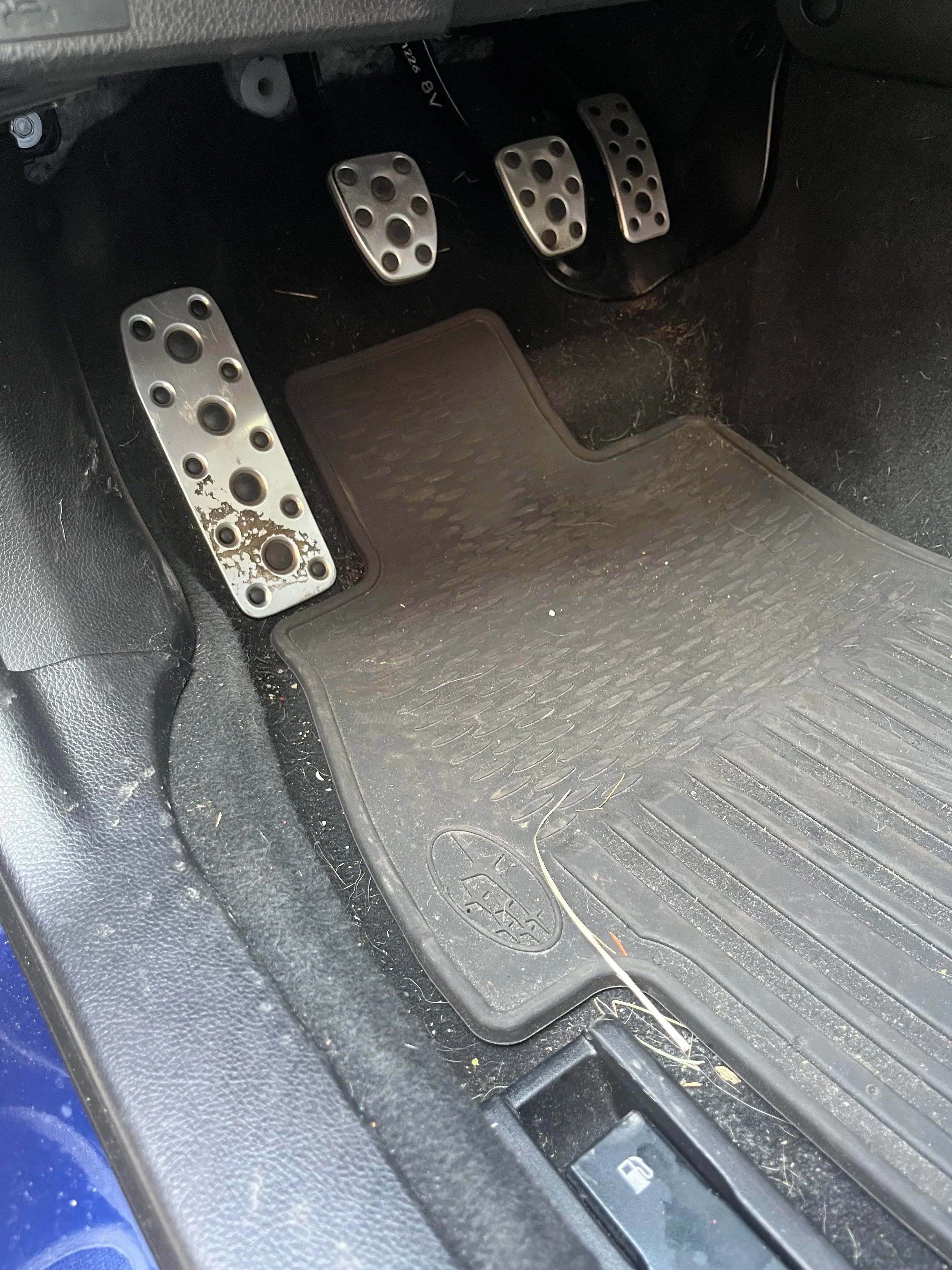 Car footwell showing four metal pedals, a rubber floor mat, and a vehicle's foot interior with some dirt and debris.