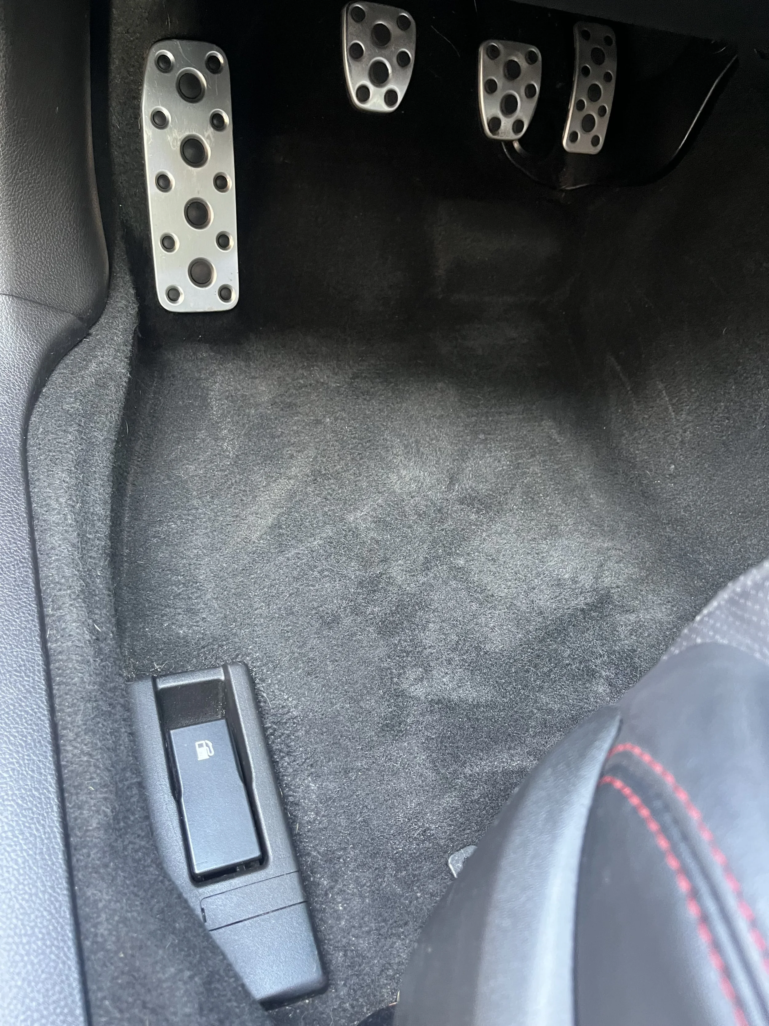 The image shows the footwell of a car with three metal pedals: clutch, brake, and accelerator, along with a small rectangular panel with a fuel icon on it.