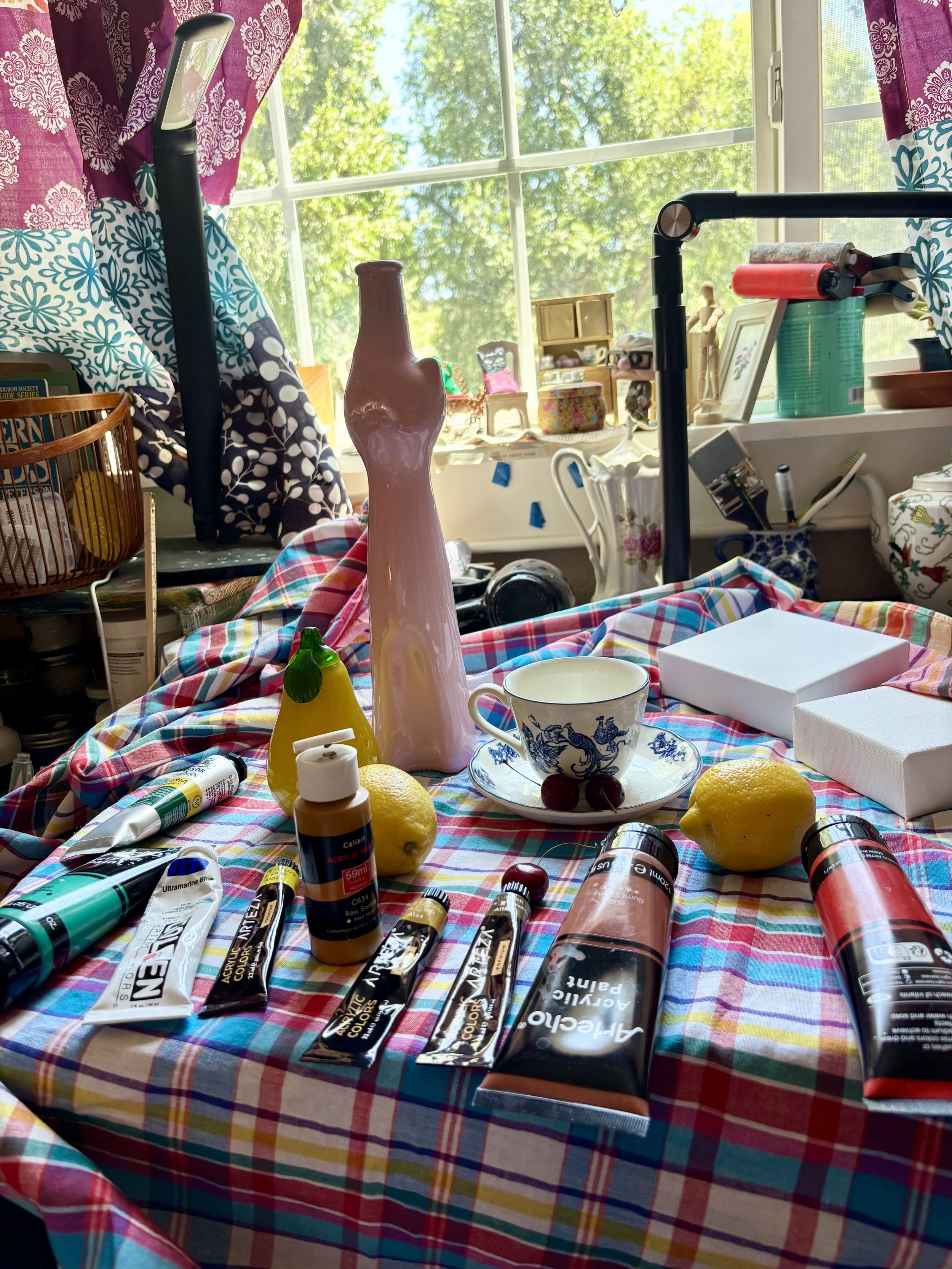 A tall pink cat shaped bottle with a teacup and saucer, two lemons, and cherries, still life on a plaid table cloth.