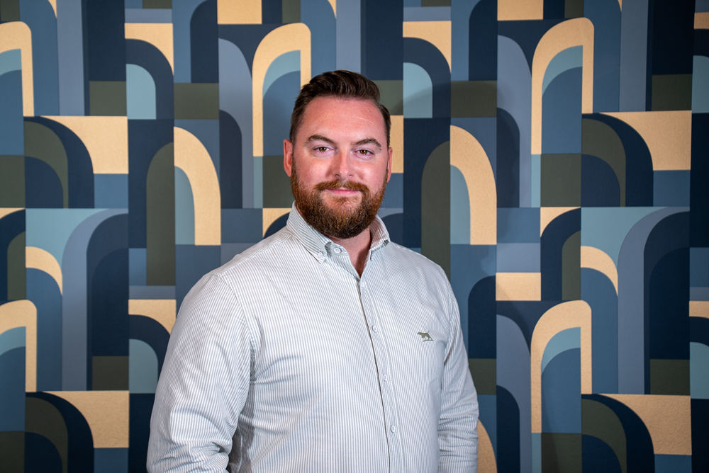 A man with a beard wearing a light gray, striped button-up shirt standing in front of a geometric patterned wall with various shades of blue and beige.