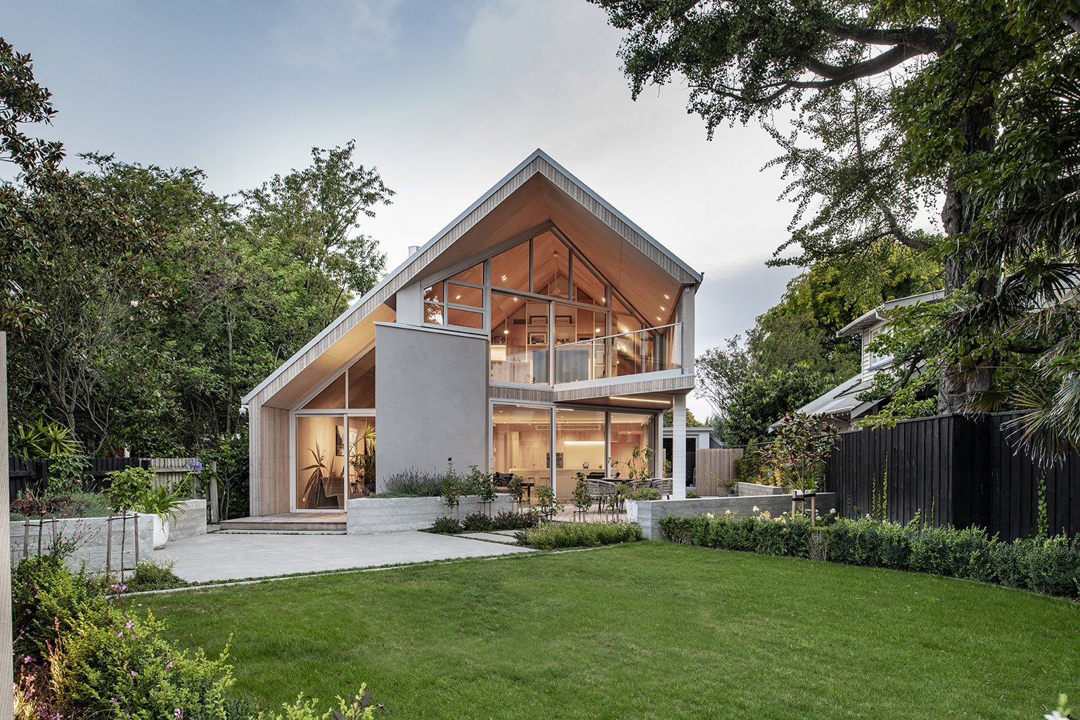 Modern two-story house with large glass windows, surrounded by greenery and a well-maintained lawn, during twilight.