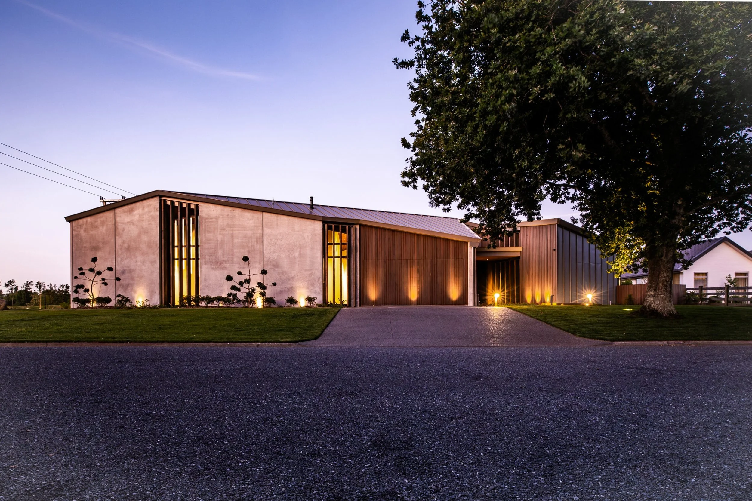 Modern house with wooden and concrete exterior, illuminated by outdoor lighting at twilight, with a large tree to the right and a well-maintained lawn.