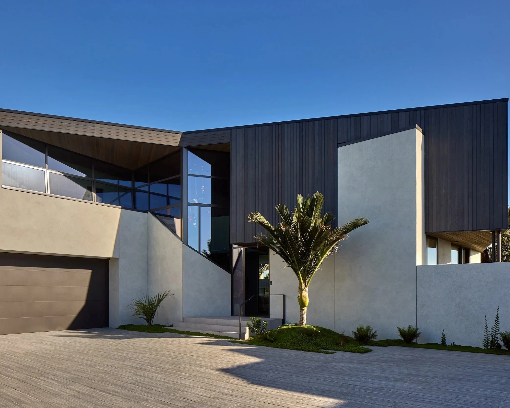 Modern house with black and beige exterior, large glass windows, palm tree, and a wooden deck under a blue sky.