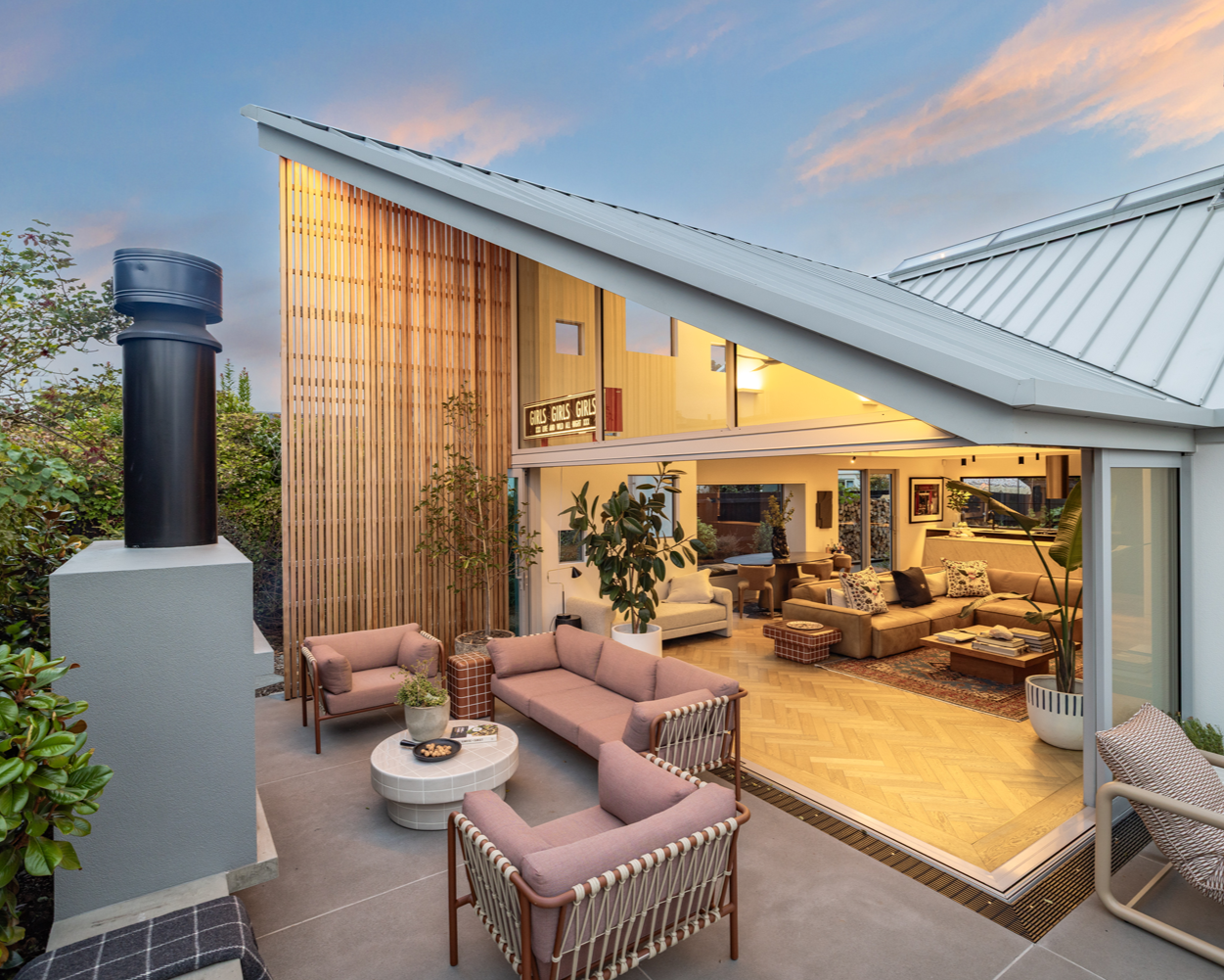 Exterior view of a modern house with a patio and interior living room visible through open sliding glass doors, featuring cozy furniture and plants, during sunset.