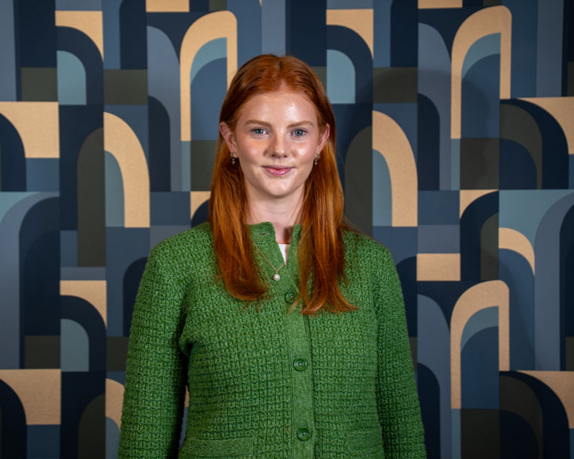 A woman with red hair wearing a green jacket standing in front of a blue and beige abstract patterned background.
