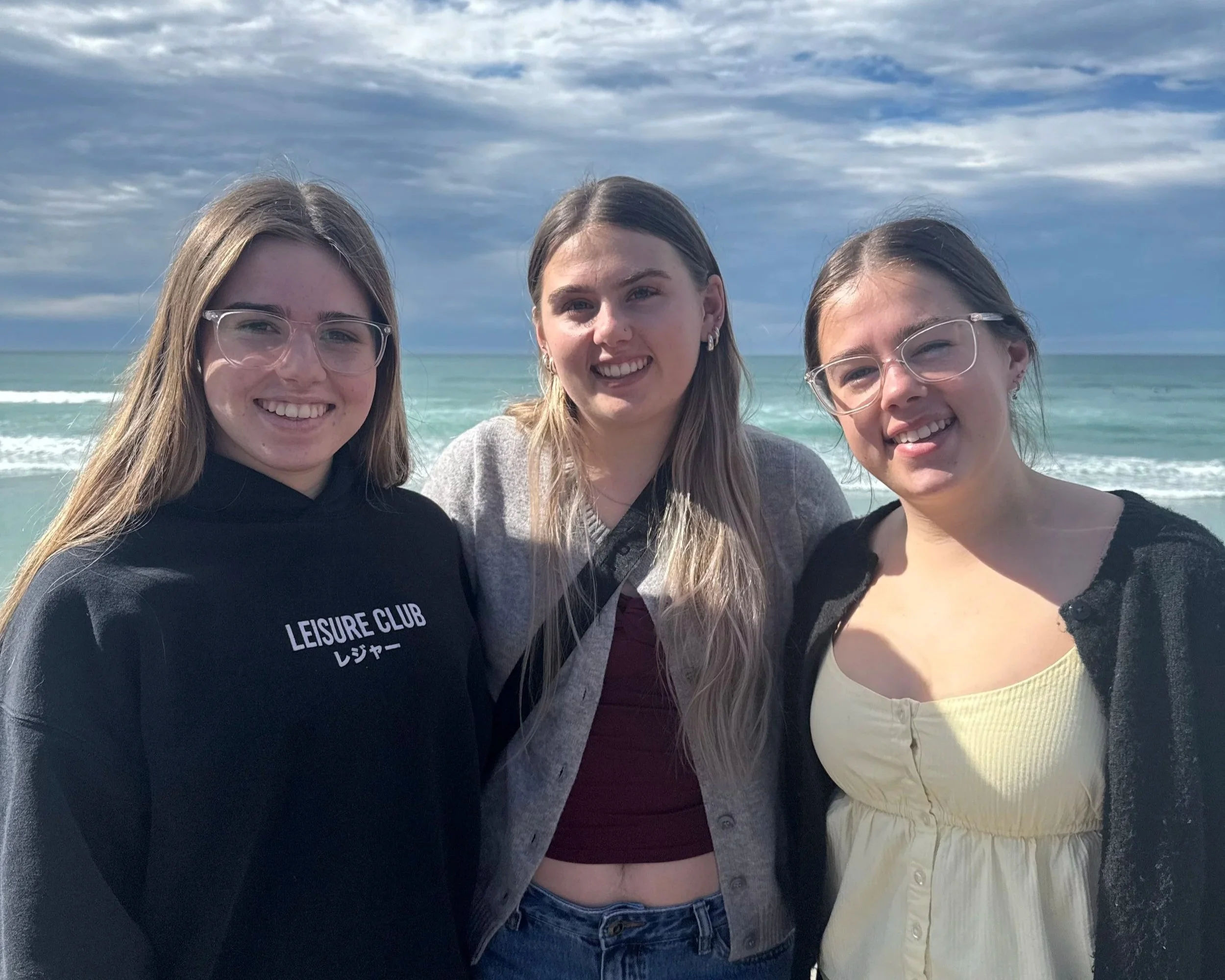 Three young women smiling at the camera on a beach with ocean and cloudy sky in the background.