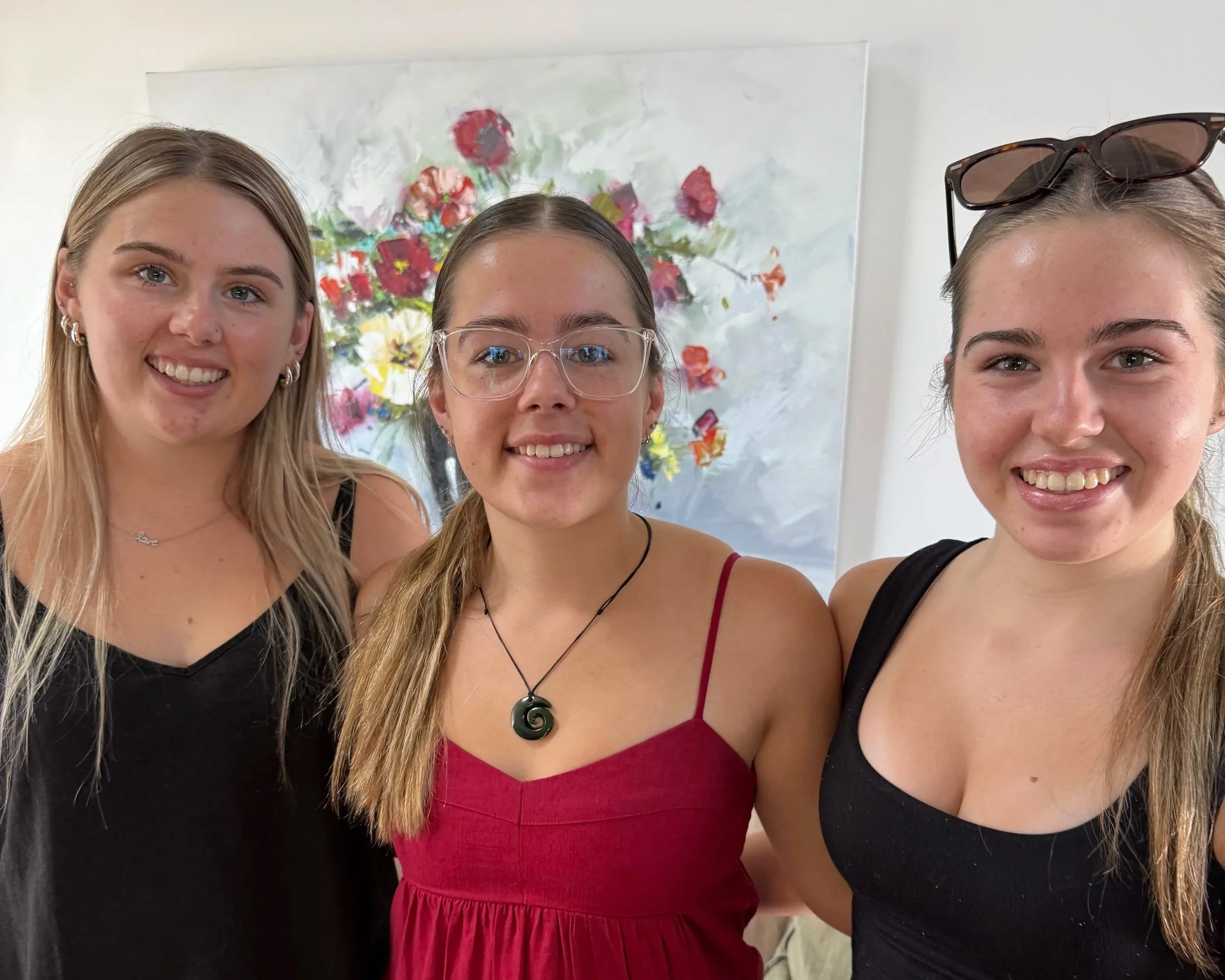 Three young women smiling in front of a colorful floral painting.