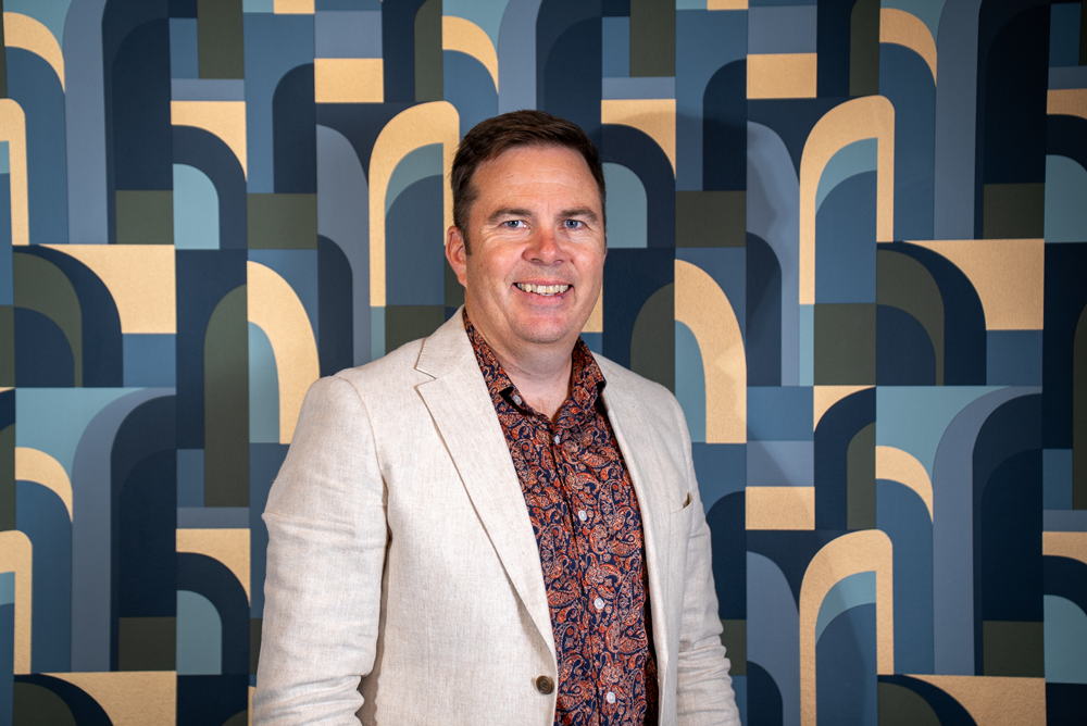 A smiling man in a beige blazer and patterned shirt stands in front of a colorful, abstract geometric background.