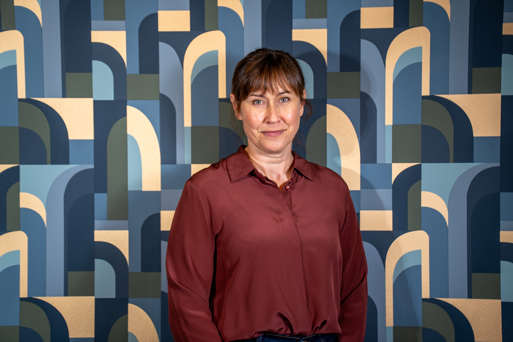 A woman with brown hair wearing a maroon satin blouse, standing in front of a colorful abstract geometric mural with arches in shades of blue, cream, and green.