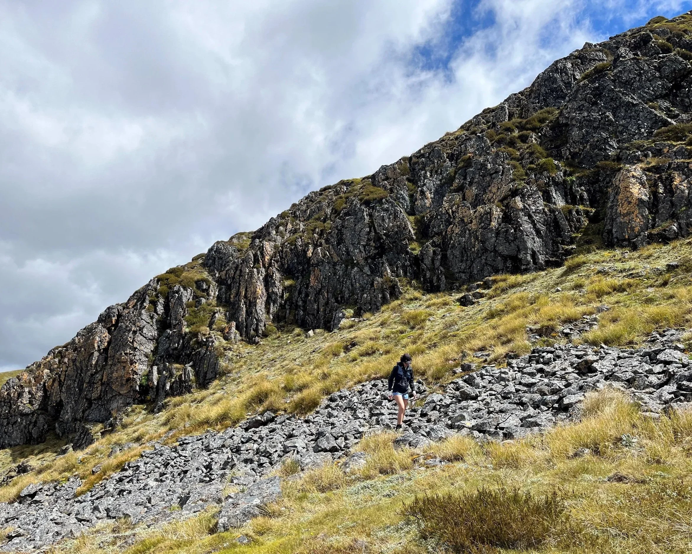 A hiker walking on a rocky slope surrounded by grass, with a steep mountain and cloudy sky in the background.