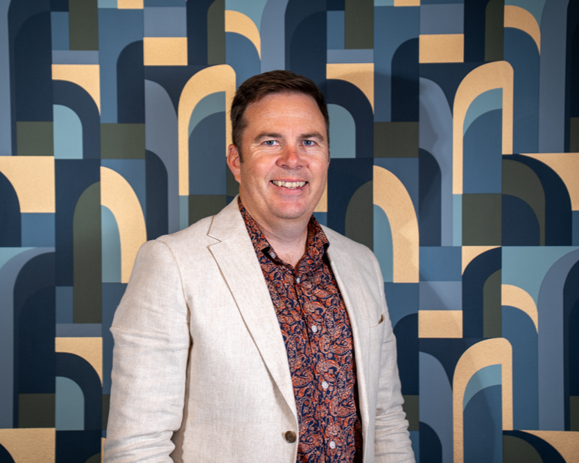 A man in a light beige blazer and patterned shirt standing in front of a geometric blue and beige abstract background.
