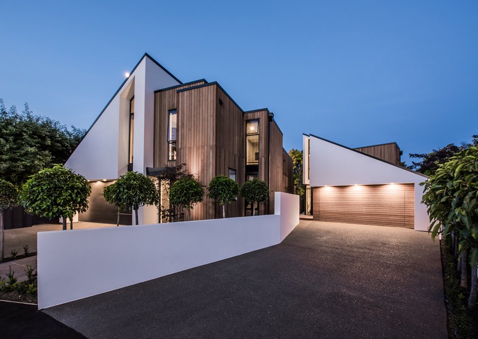 Modern house with white and wooden exterior, trees along the driveway, and a garage with a wooden door, during dusk.