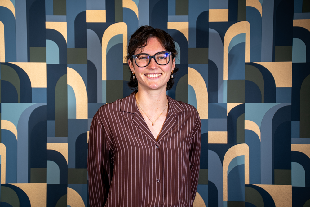 A woman with short dark hair, glasses, and earrings, smiling in front of a geometric patterned wall with arches in various shades of blue, beige, and green.