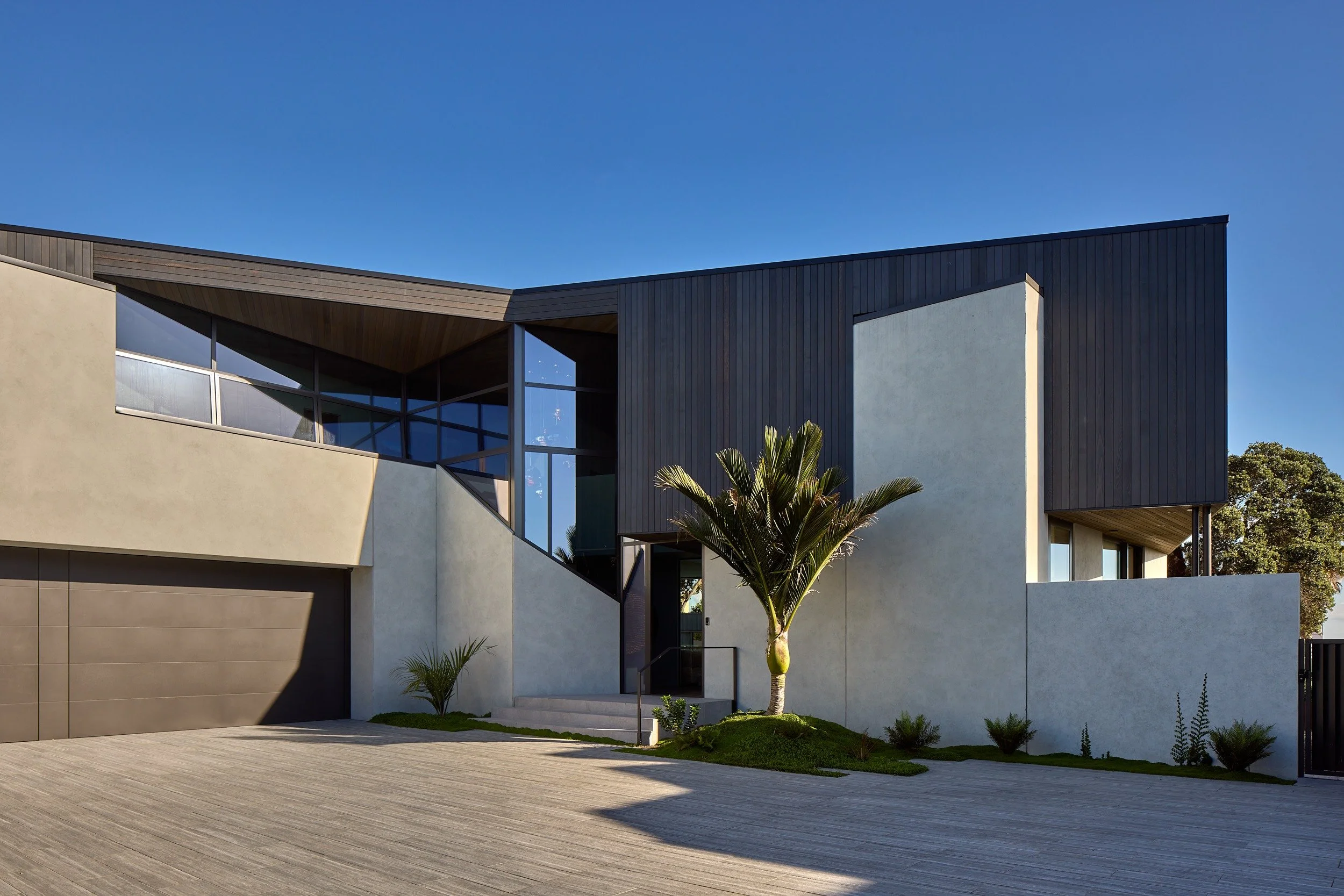 Modern house with concrete and dark wood exterior, large glass windows, and landscaping including palm trees under a clear blue sky.
