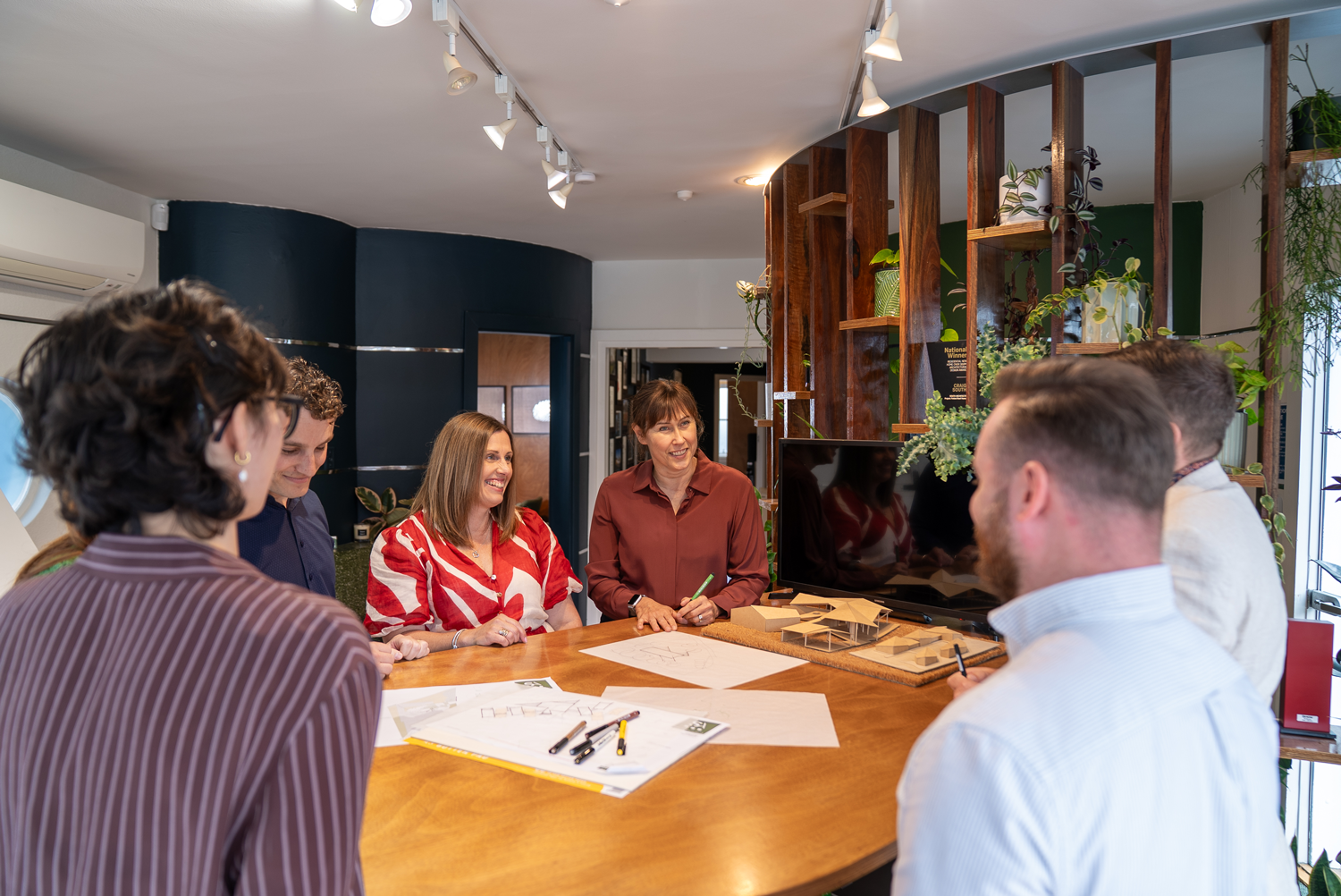 A group of six people gathered around a wooden conference table having a discussion. There are papers, markers, and architectural models on the table. A woman in a burgundy shirt is talking and smiling, while others listen and participate. The room has indoor plants and modern decor.