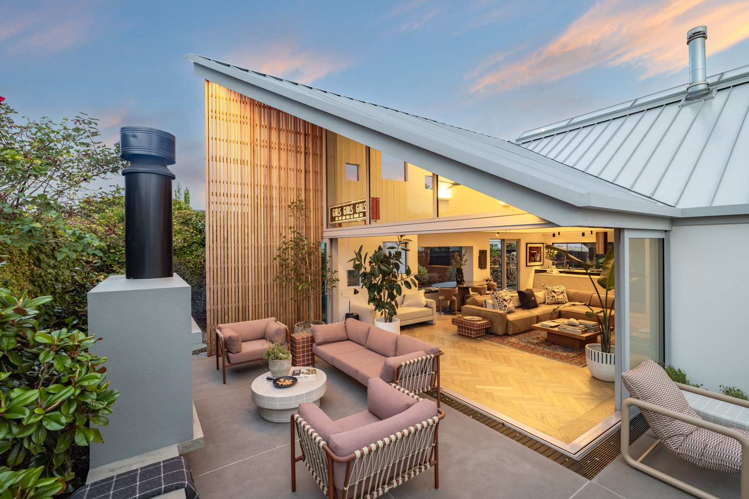 Modern outdoor patio with pink sofas, chairs, and a coffee table, leading into a stylish living room with beige sofas, potted plants, and abstract art, during sunset.